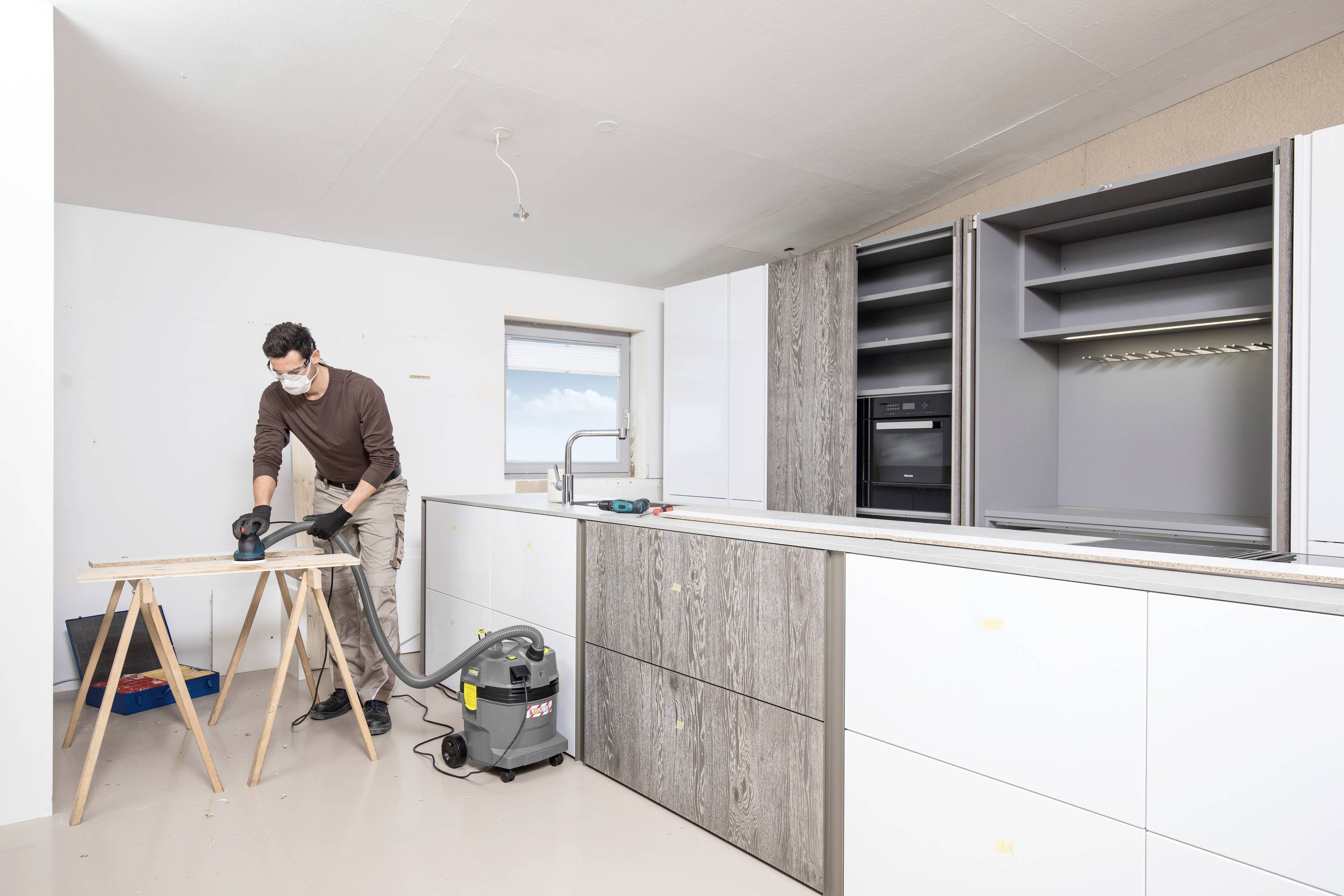 Ein Mann arbeitet in einer hellen Küche an einem Holzbrett. Er trägt eine Schutzmaske und Handschuhe. Neben ihm steht ein Staubsauger.