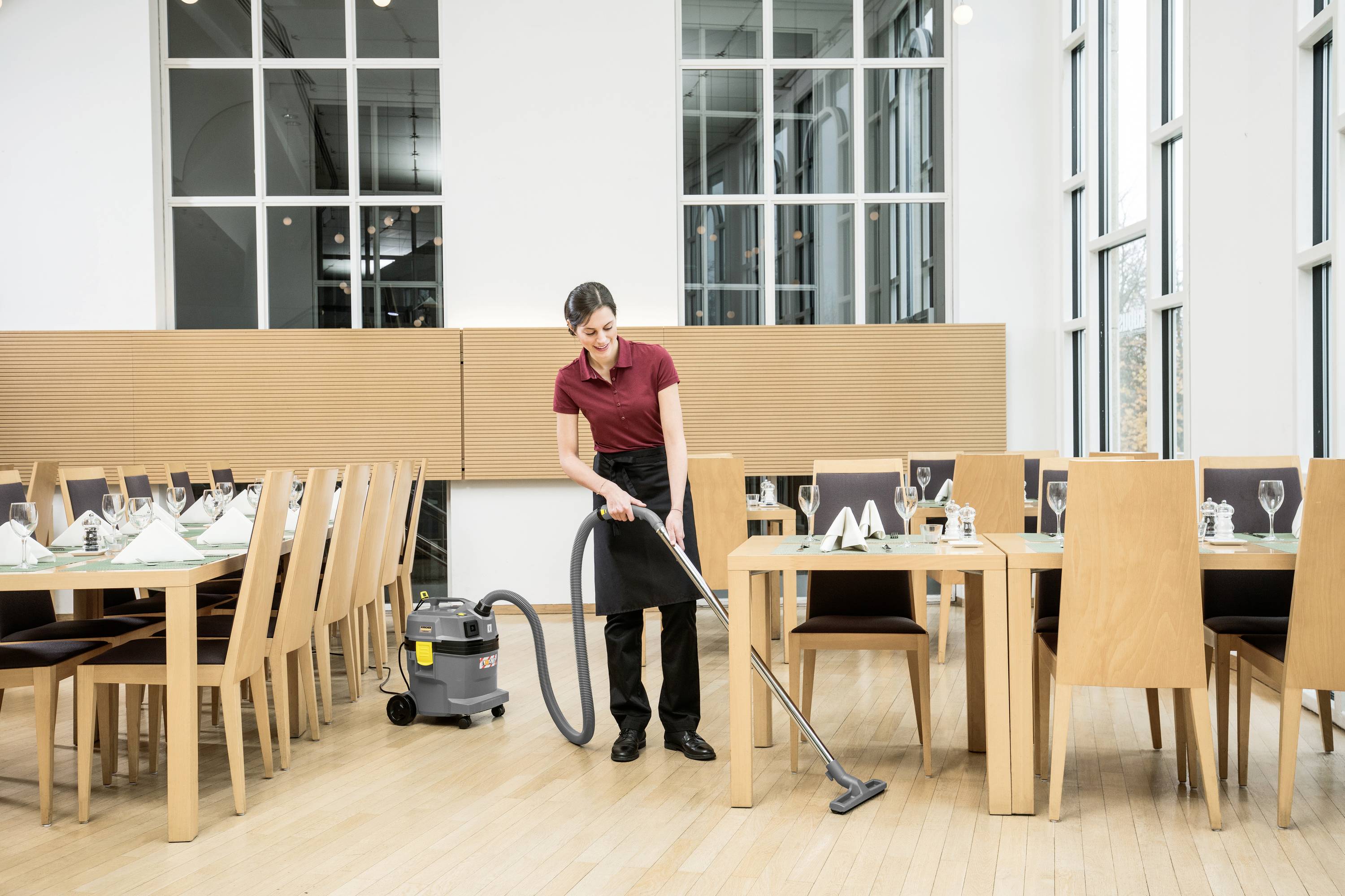Eine Person saugt den Boden in einem leeren Restaurant mit einem Tischstaubsauger. Tische sind gedeckt, große Fenster im Hintergrund.