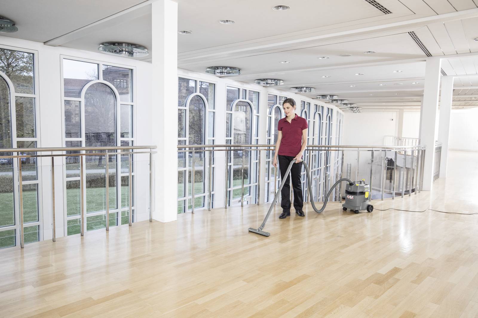 Eine Person in einem roten Hemd benutzt einen Staubsauger auf einem polierten Holzboden in einem geräumigen, modernen Gebäude mit großen Fenstern.