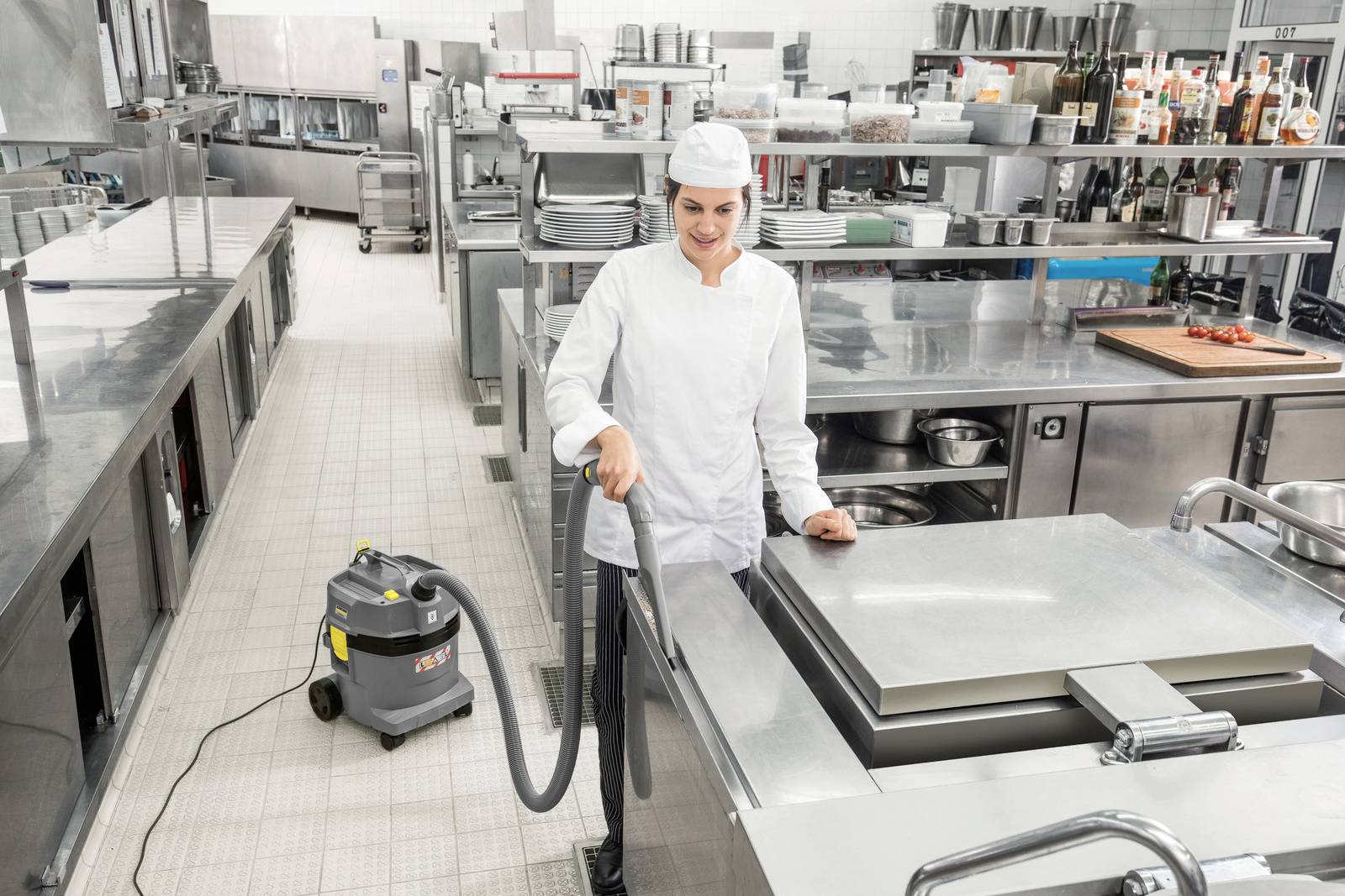 Ein Koch in einer gewerblichen Küche verwendet einen Staubsauger, um Edelstahloberflächen zu reinigen, umgeben von industriellen Geräten und Kochgeschirr.