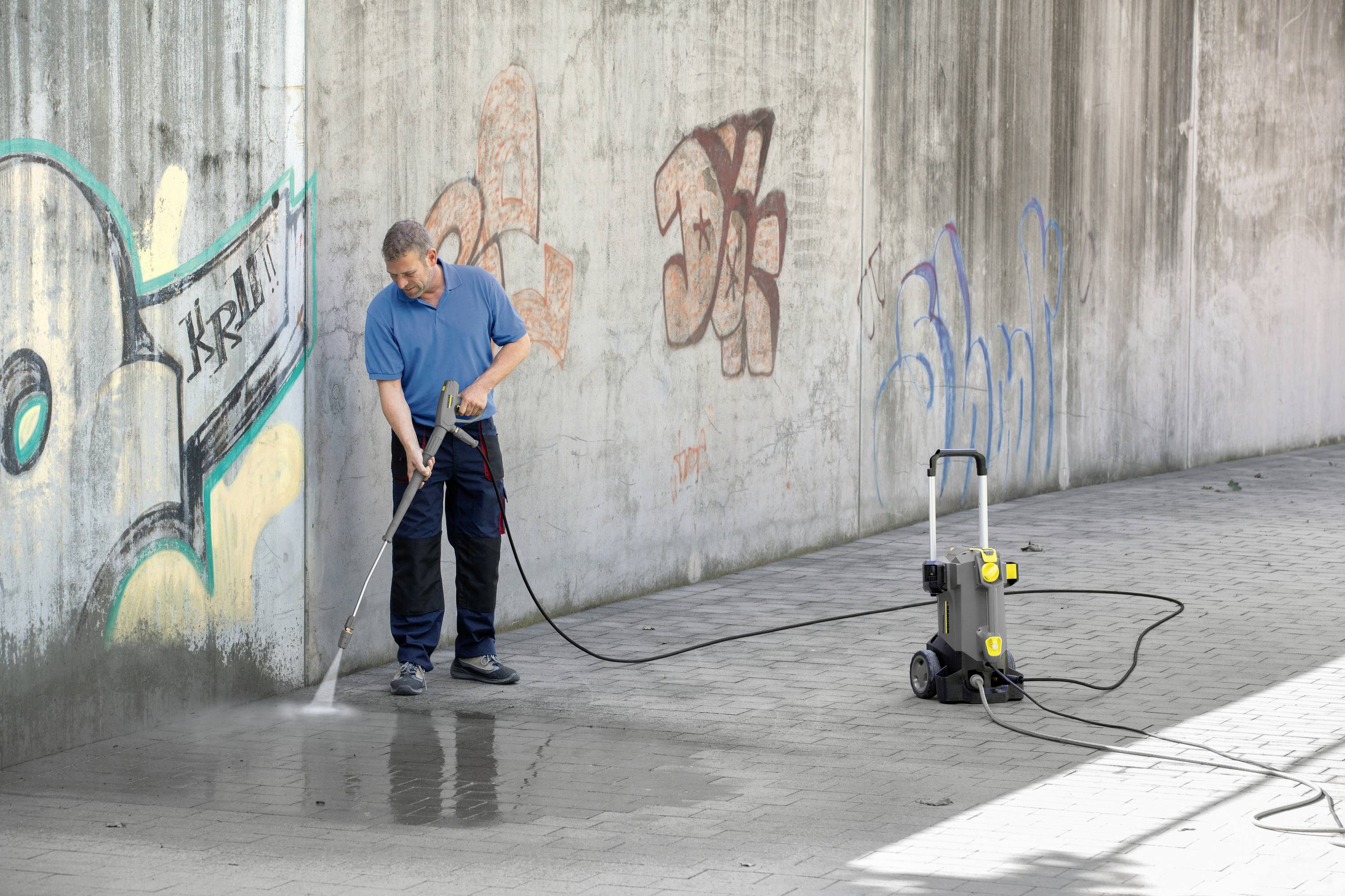 Eine Person reinigt eine mit Graffiti bedeckte Mauer mit einem Hochdruckreiniger auf einem gepflasterten Boden.