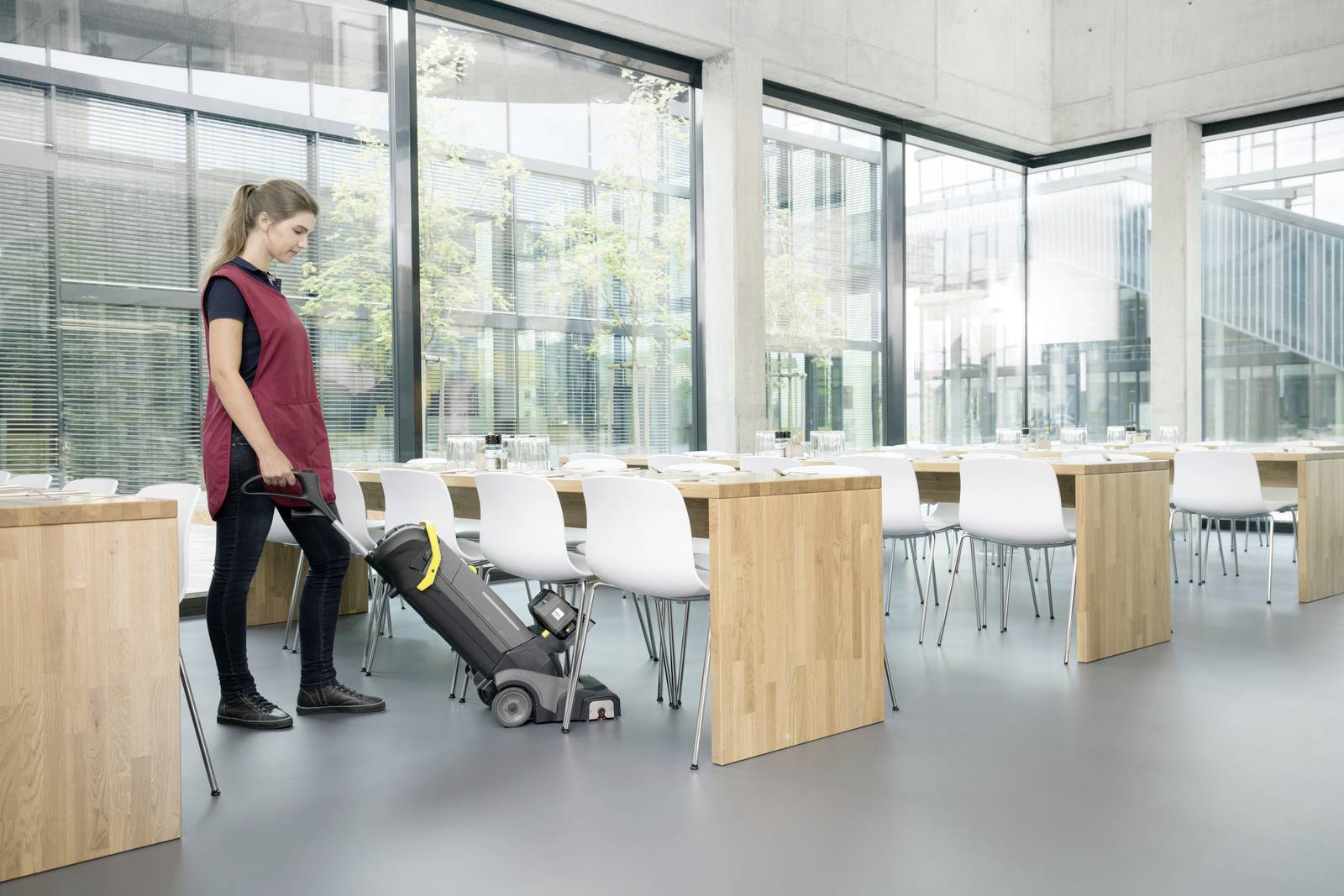 Eine Person reinigt den Boden eines leeren, modernen Konferenzraums mit einem Staubsauger. Tische und Stühle sind ordentlich angeordnet.