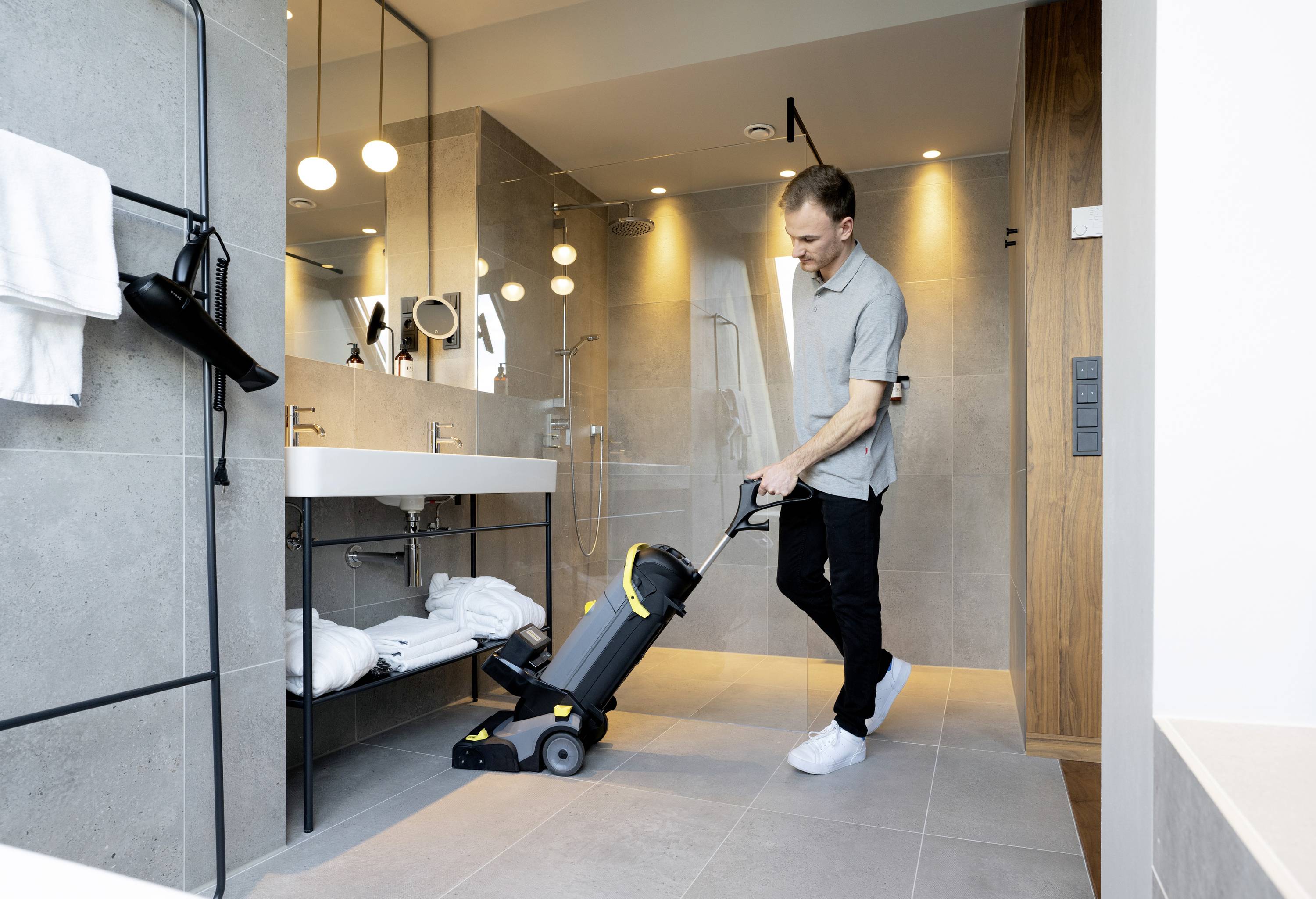 Ein Mann benutzt eine Bodenreinigungsmaschine in einem modernen Badezimmer mit Dusche, großen Spiegeln und Handtüchern auf dem Regal.