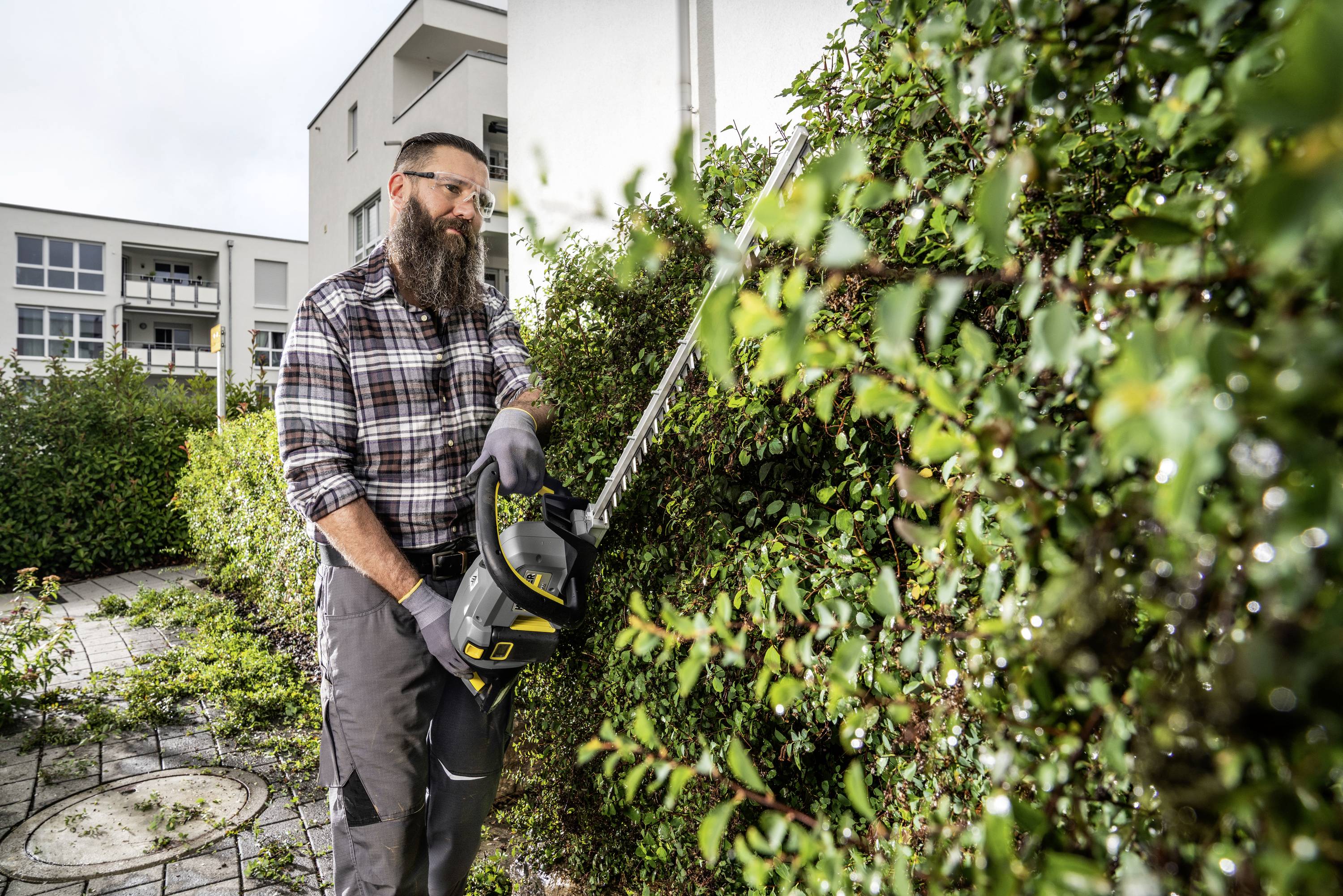Ein Mann mit Bart trimmt eine Hecke mit einer elektrischen Heckenschere in einem modernen Wohngebiet.