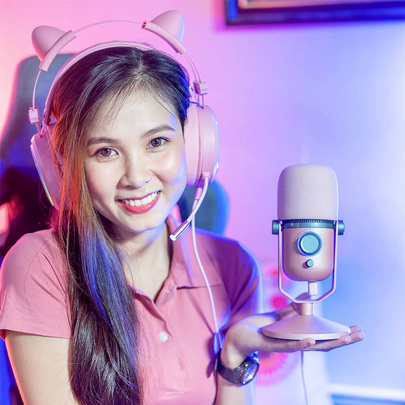 Eine lächelnde Person sitzt vor einem Mikrofon und trägt Kopfhörer mit Katzenohren. Sie trägt ein rosa Polohemd.