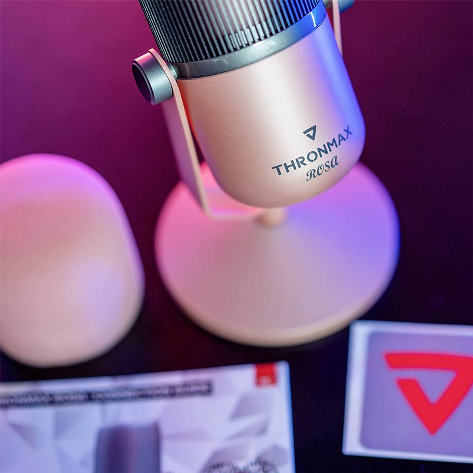 Ein rosafarbenes Mikrofon mit der Aufschrift 'Thronmax Rosa' steht auf einem Tisch. Daneben befindet sich ein runder Gegenstand und ein Heft mit einem Logo.