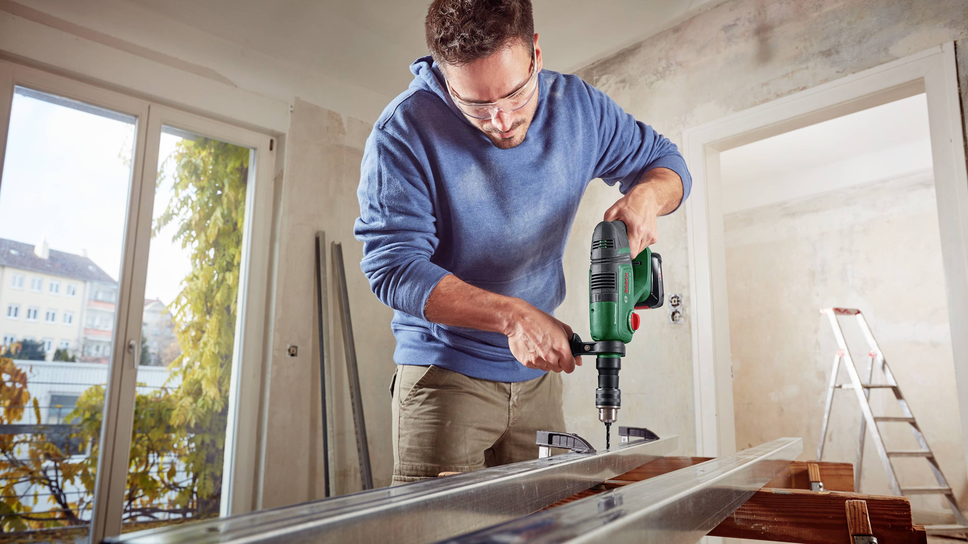 Ein Mann in Schutzbrille und blauem Hoodie benutzt eine grüne Bohrmaschine, um ein Loch in einen Metallträger in einem renovierten Raum zu bohren.