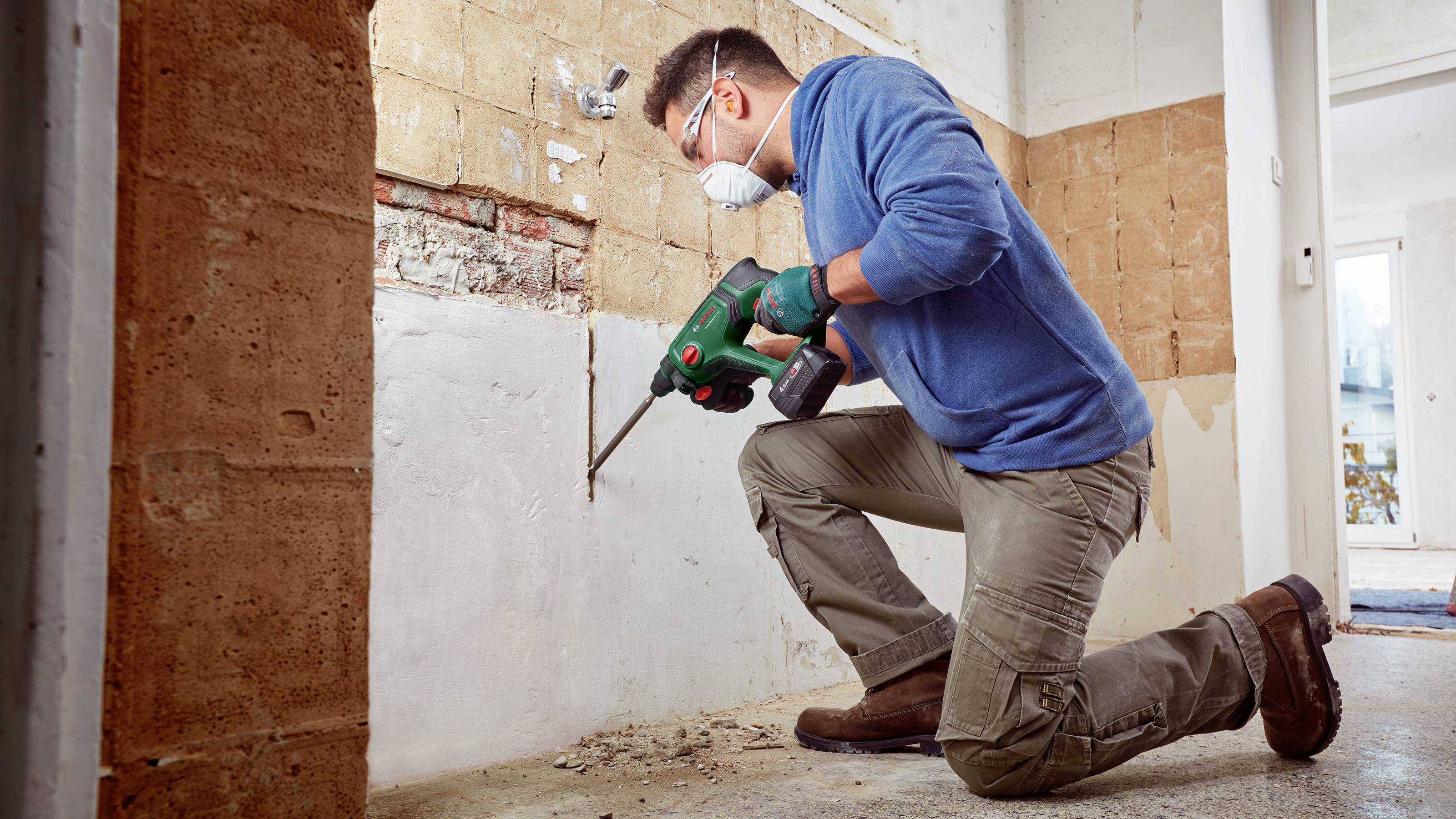 Ein Mann mit Schutzbrille und Maske renoviert eine Wand mit einem Bohrhammer. Er trägt Handschuhe und kniet auf einem grauen Boden.