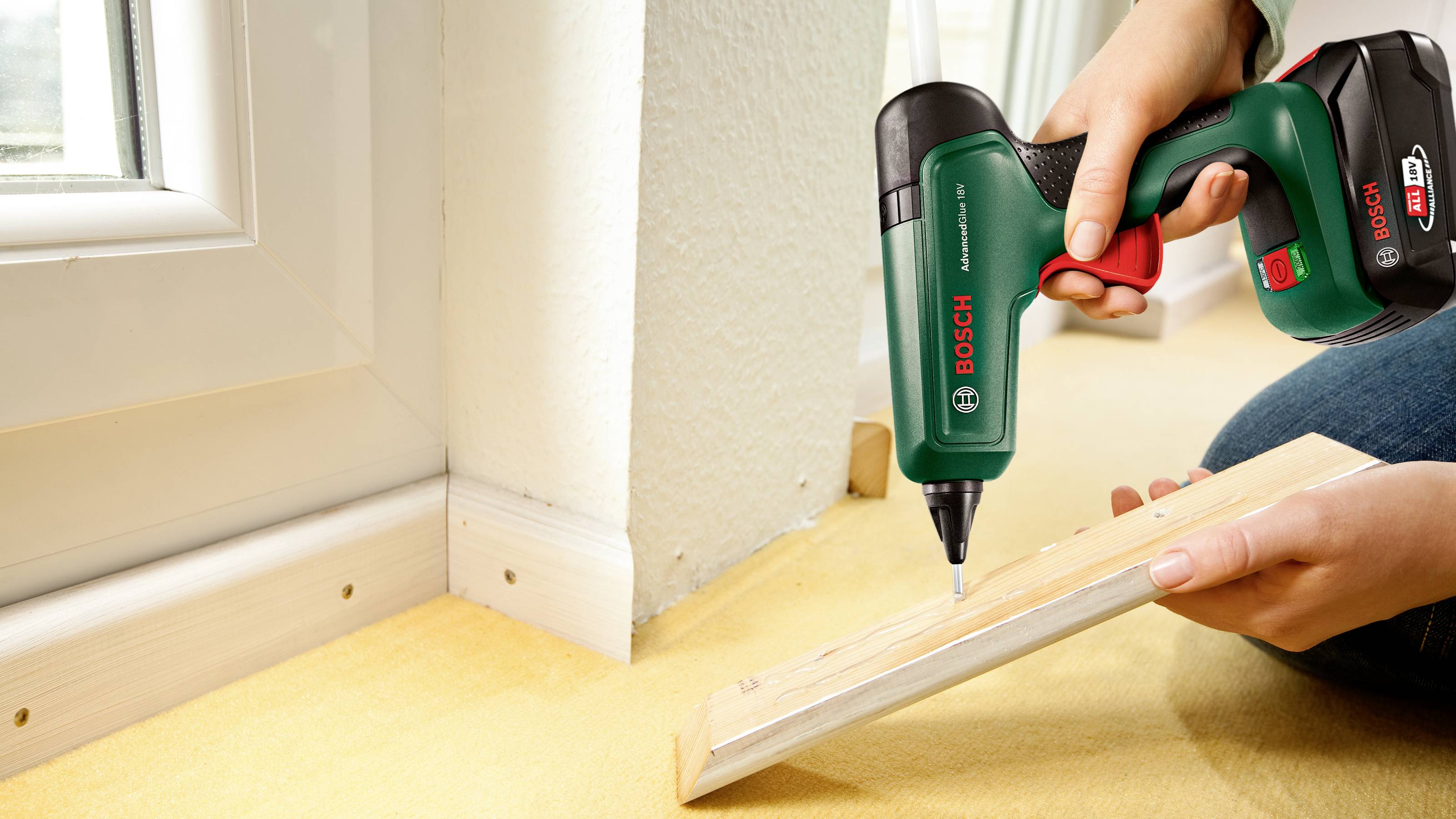 Eine Person verwendet einen Bosch-Akkuschrauber, um eine Holzleiste in der Ecke eines Raumes neben einem Fensterrahmen zu befestigen.
