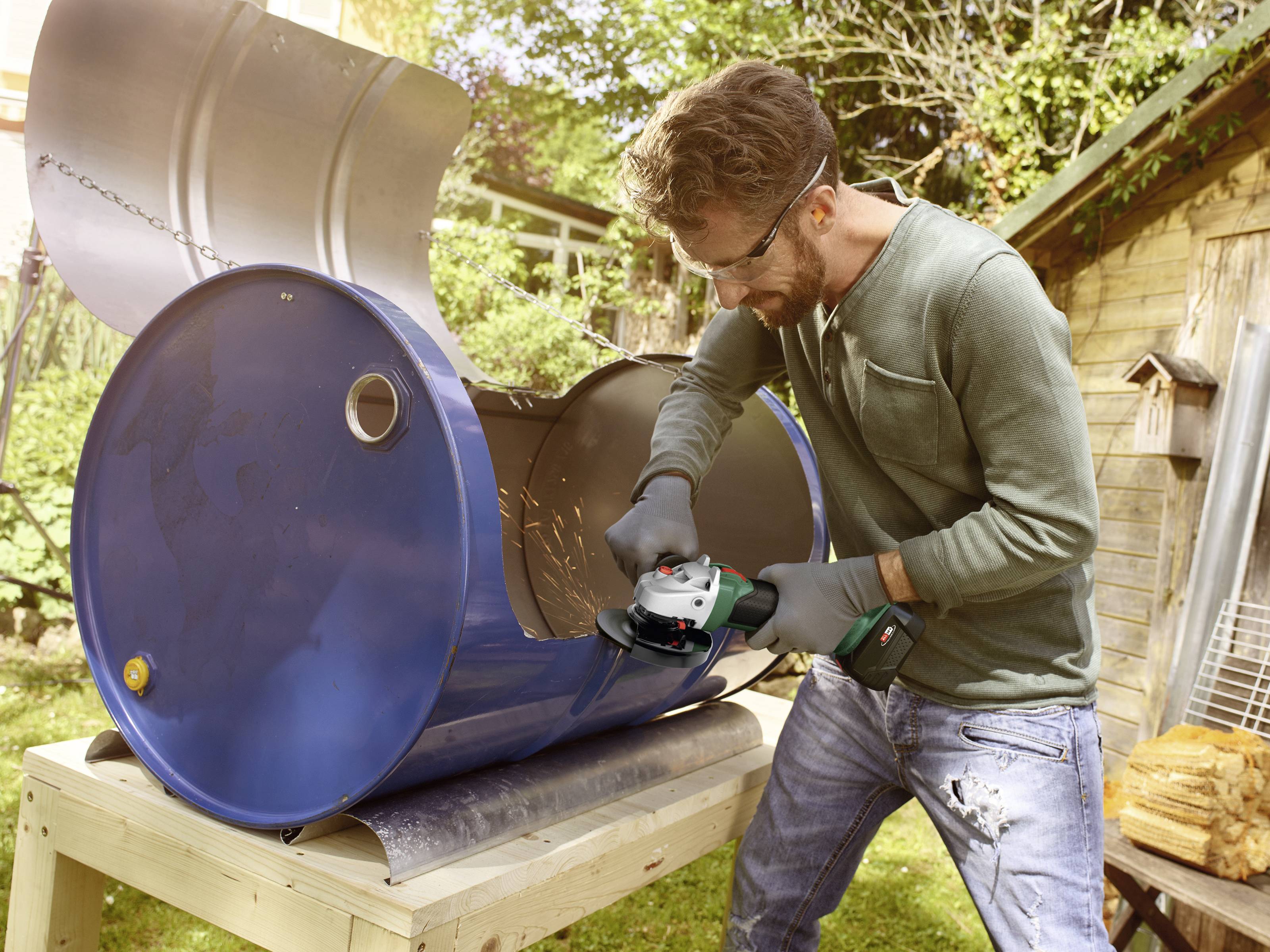 Ein Mann mit Schutzbrille und Handschuhen schneidet mit einem Winkelschleifer ein großes, liegendes Metallfass im Garten auf.