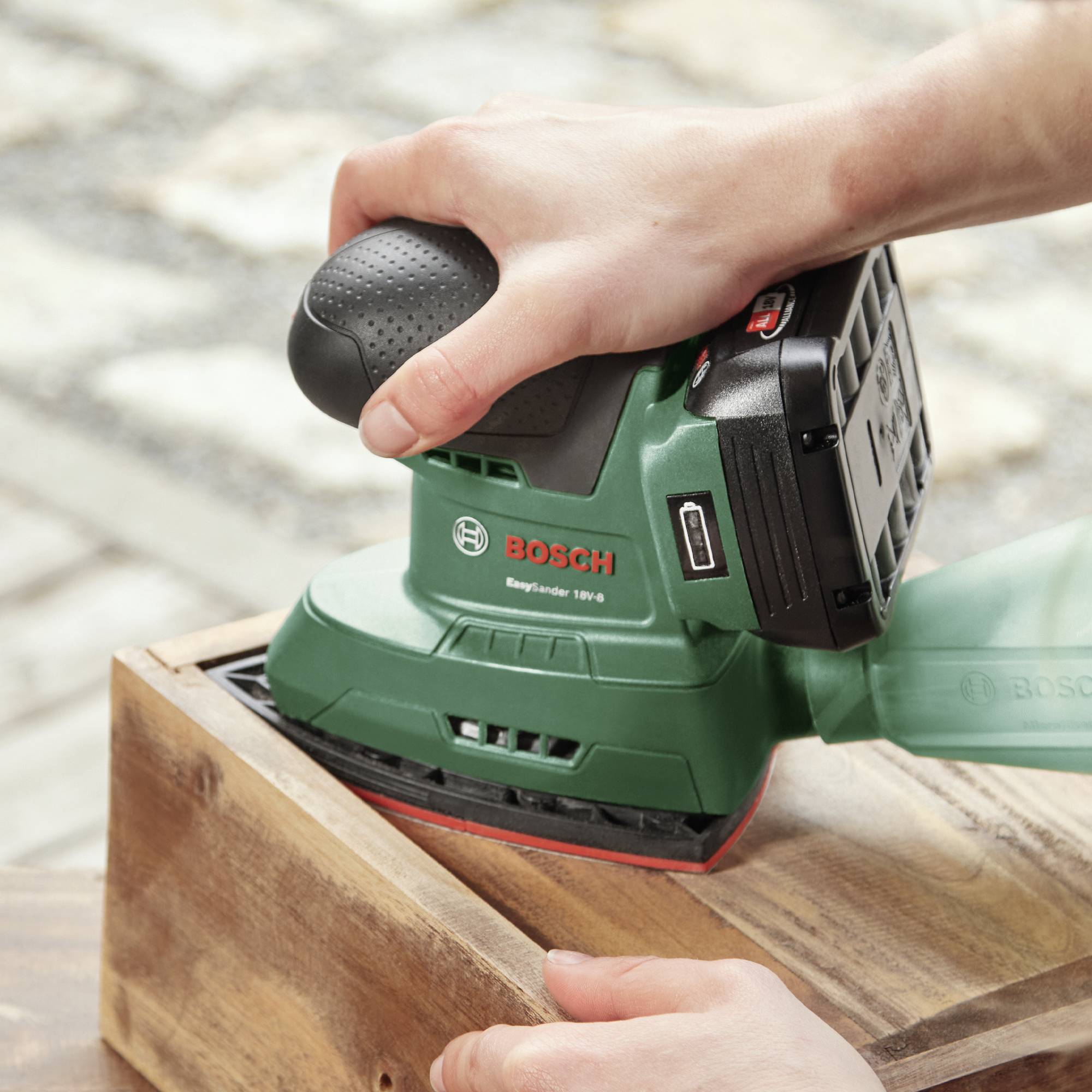 Eine Person schleift ein Holzstück mit einem grünen Bosch-Schleifgerät. Die Hand hält das Werkzeug sicher, während es auf dem Holz bewegt wird.