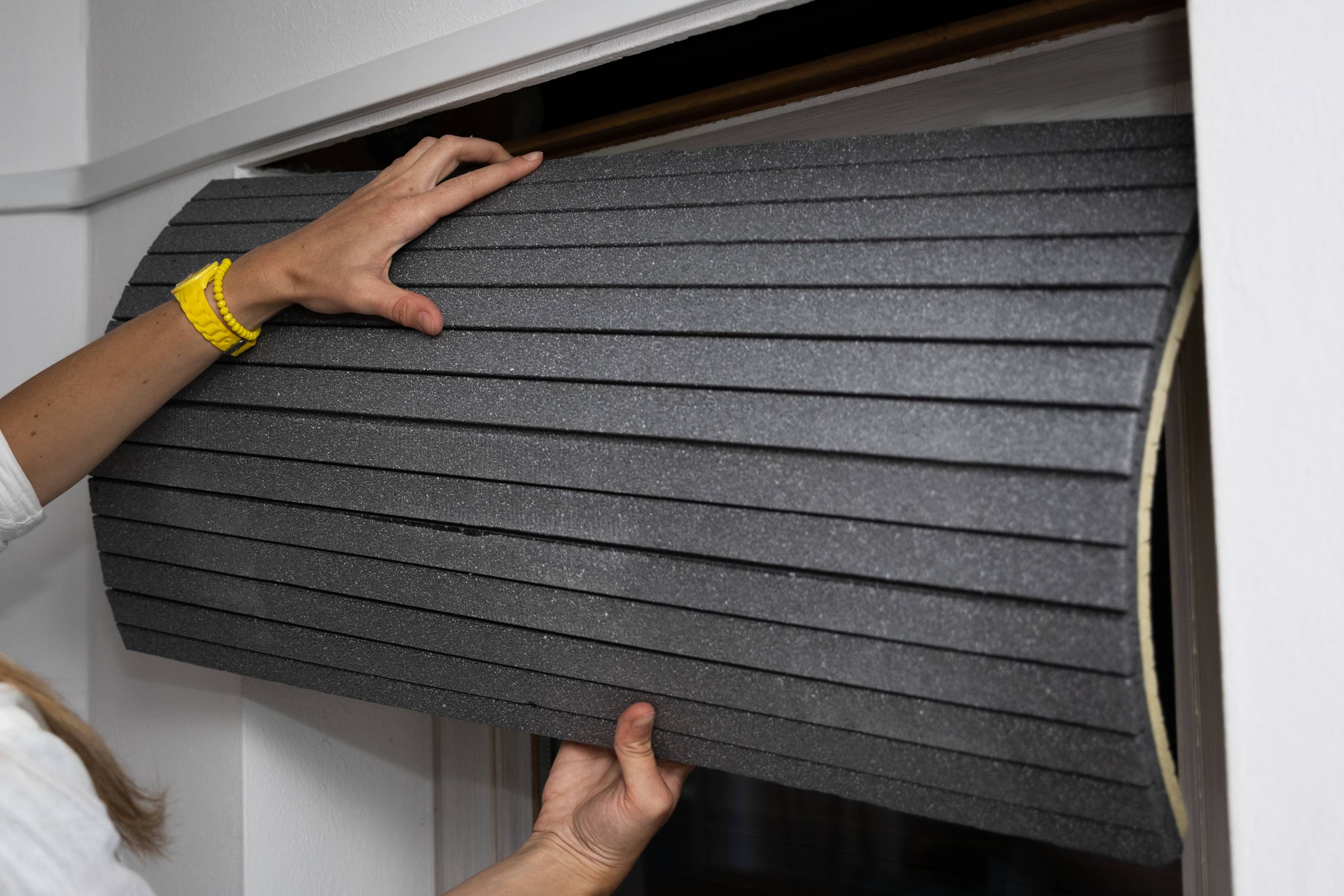 Eine Person installiert eine gerollte, schwarze Schaumstoff-Dämmmatte in einen Fensterrahmen. Hände halten die Matte an beiden Enden.