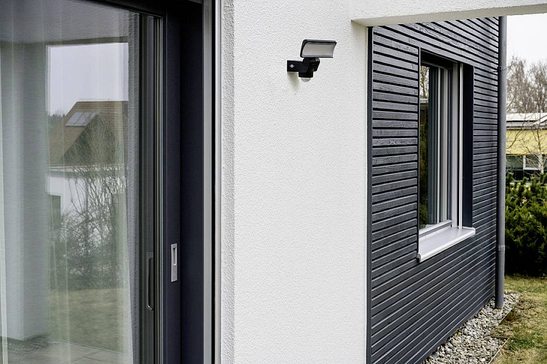 Ein modernes Haus mit weißen und grauen Wänden; ein Bewegungsmelder ist neben einem Fenster und einer Glastür angebracht.