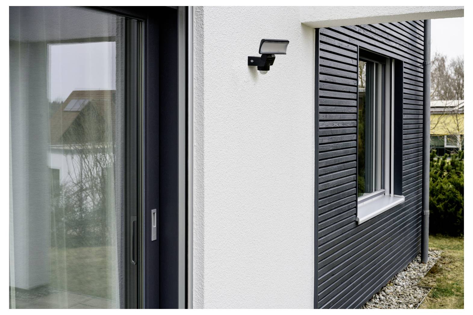 Außenansicht eines modernen Hauses mit einer Glasschiebetür, einer wandmontierten Leuchte und dunkler Verkleidung neben einem Fenster mit Kieslandschaftsbau.