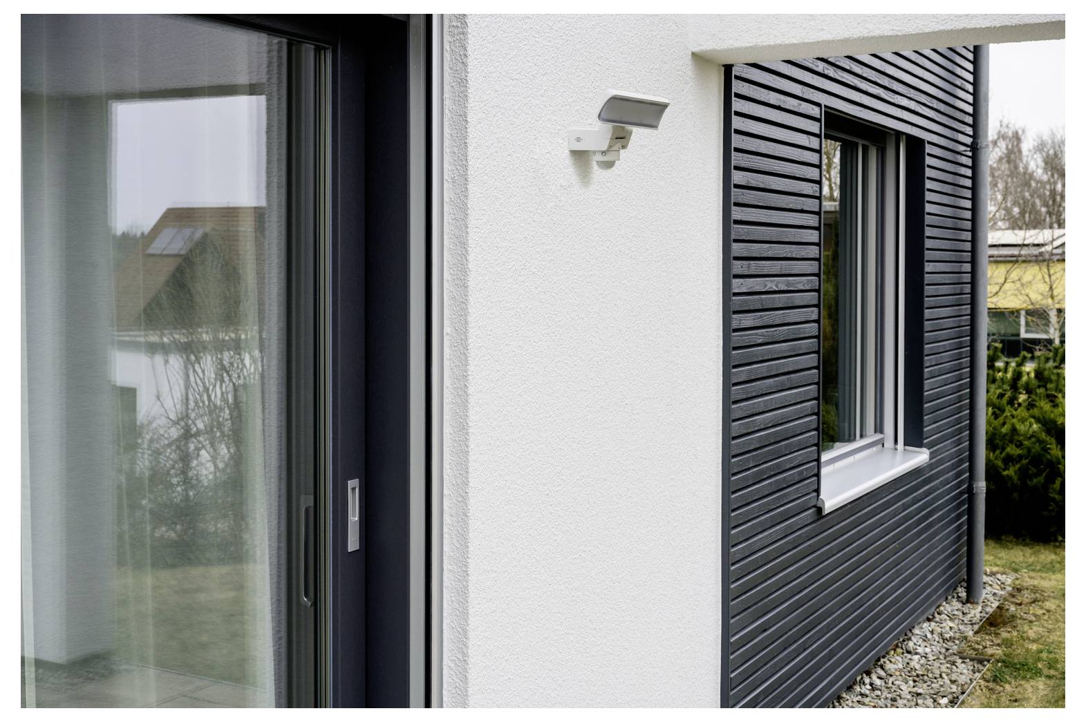 Außenansicht eines modernen Hauses mit einer Glasschiebetür, weißer Stuckwand mit Wandleuchte und dunkler horizontaler Verschalung neben einem Fenster.