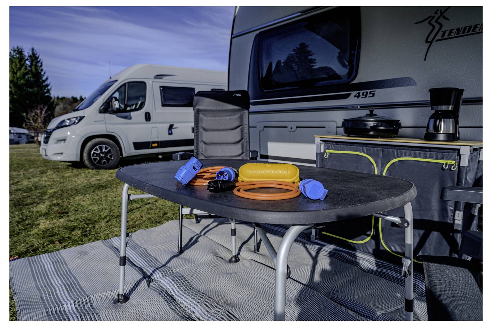 Campingplatz neben zwei Wohnmobilen, mit einem Outdoor-Klapptisch mit Schläuchen und Campingausrüstung auf einer gestreiften Matte.