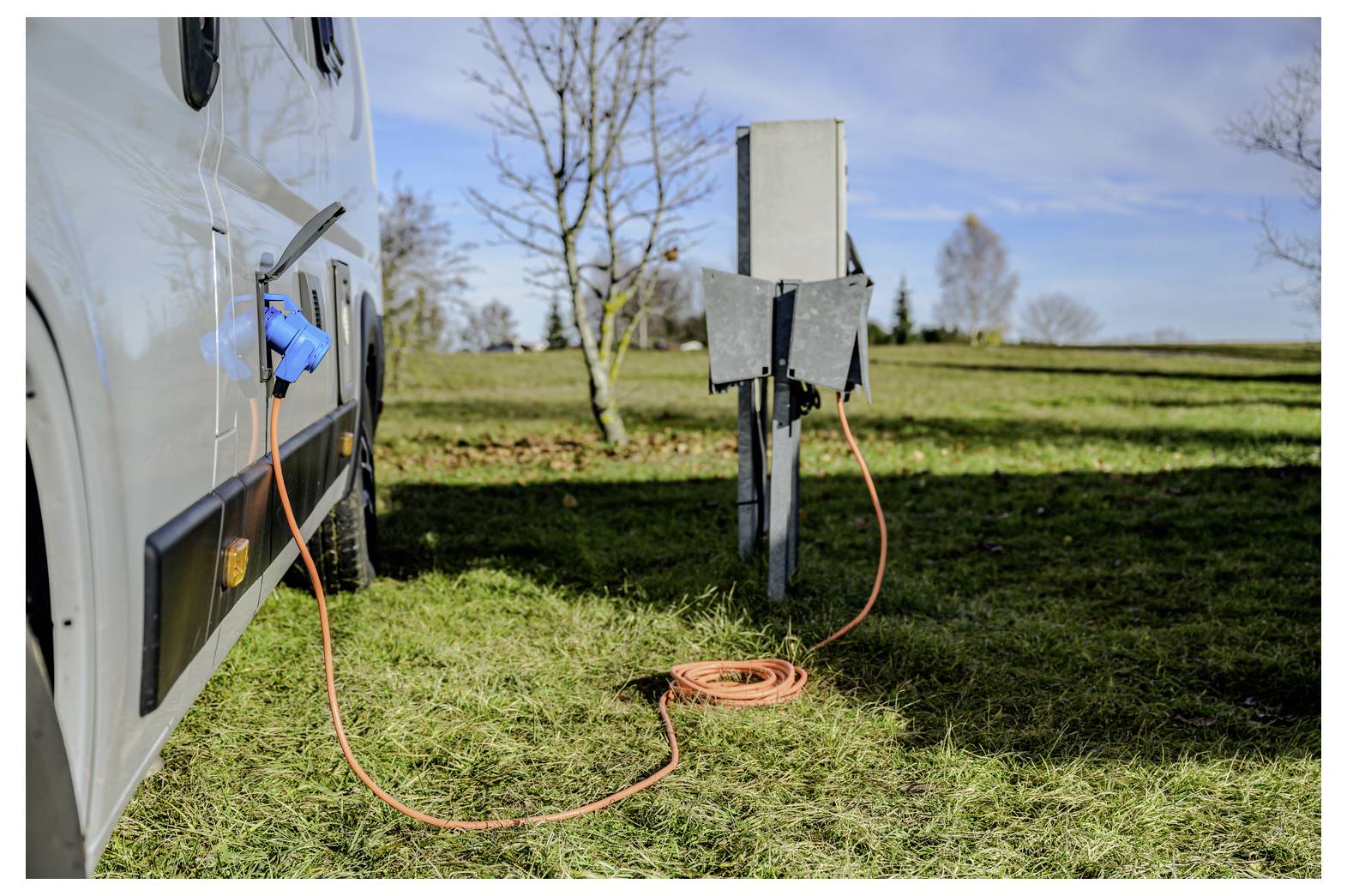 Wohnmobil, das mit einem orangefarbenen Verlängerungskabel an einen Strompedestal auf einem Campingplatz angeschlossen ist, auf einer Grünfläche