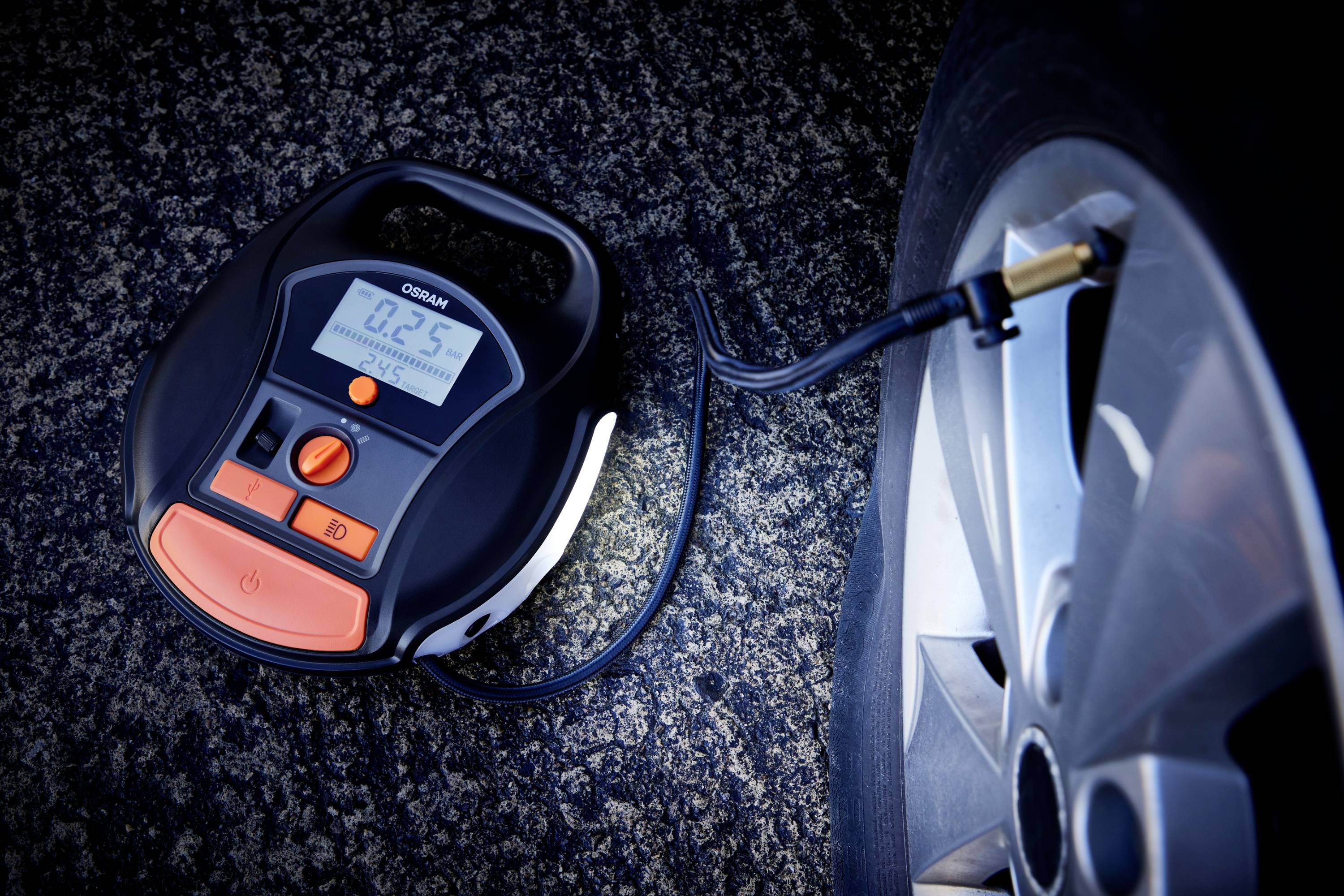 Ein digitaler Reifenkompressor pumpt einen Autoreifen auf. Der Bildschirm zeigt '0.25' Bar an. Eine Straße bildet den Hintergrund.