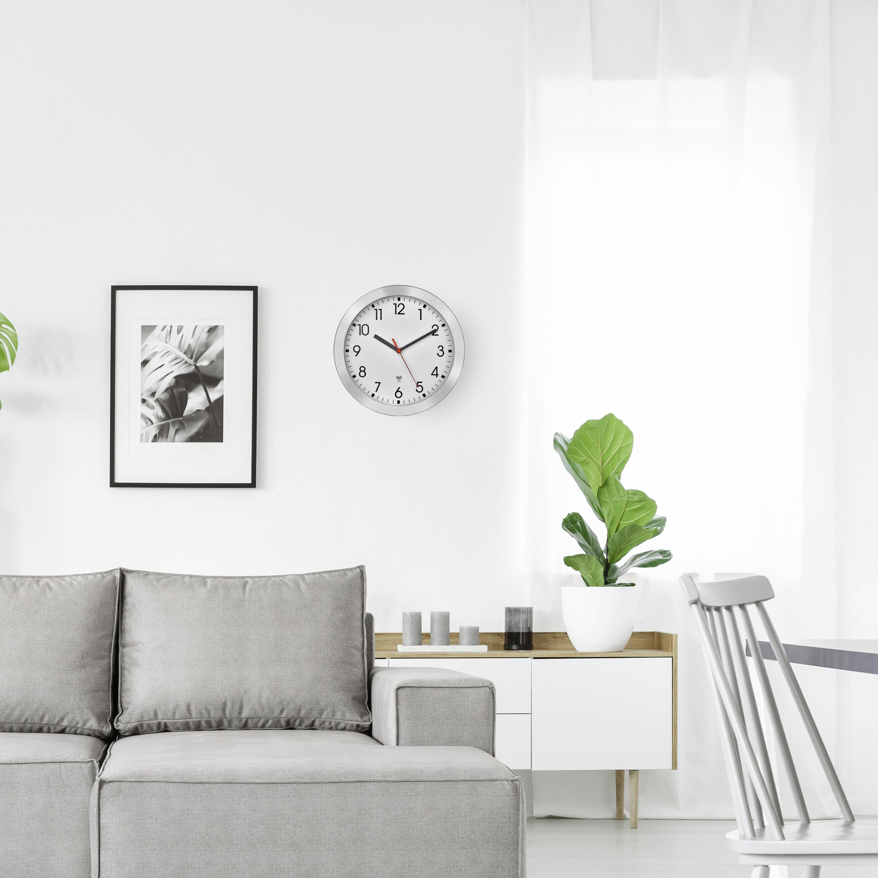 Ein modernes Wohnzimmer mit grauem Sofa, Uhr an der Wand, weißem Regal und Zimmerpflanzen. Helle, minimalistische Einrichtung.