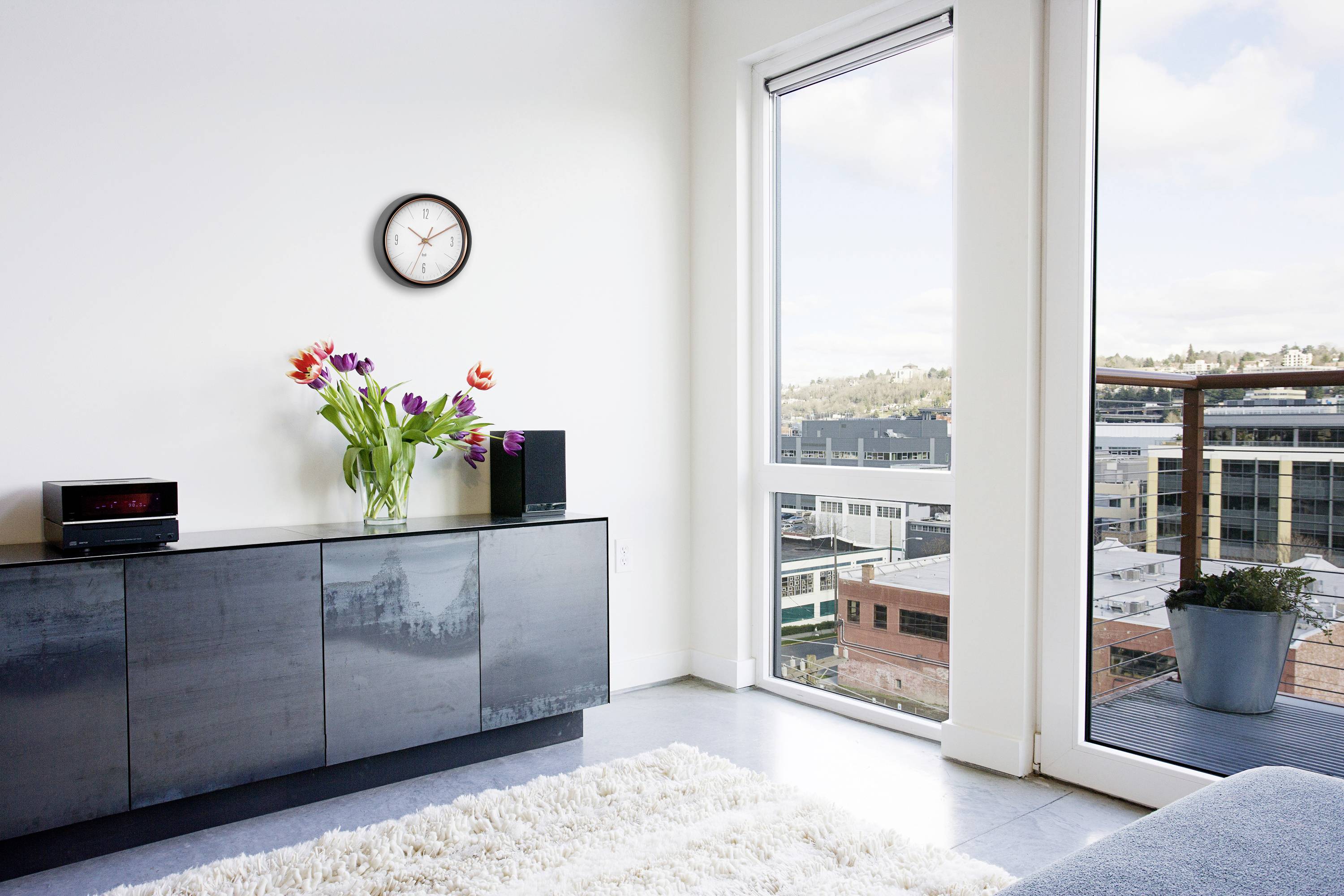 Moderne Wohnzimmeransicht mit klaren Fensterblick auf Stadtgebäude, Uhr an der Wand, Sideboard mit Vase bunter Blumen.