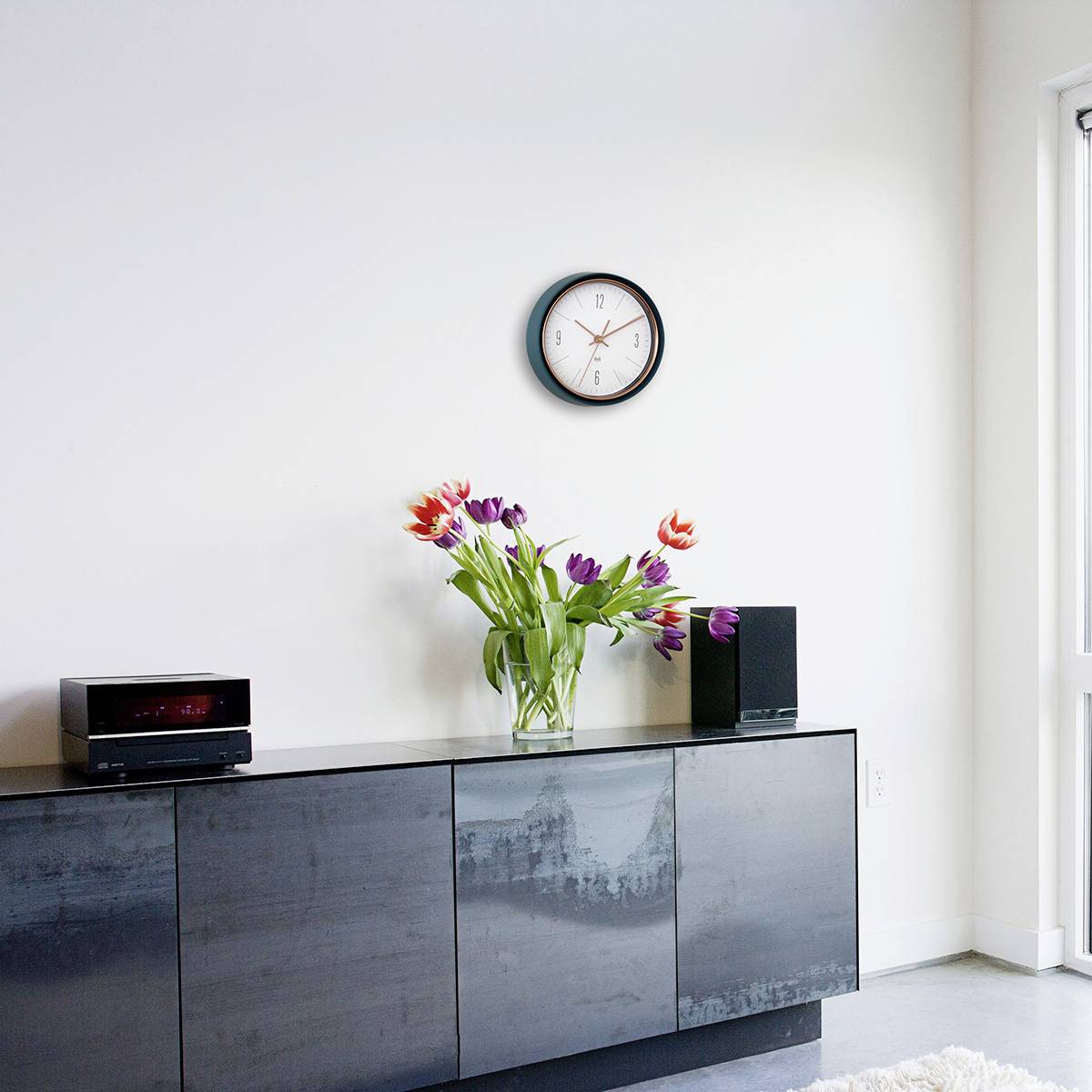 Ein modernes Wohnzimmer mit einem grauen Sideboard, einer Vase mit bunten Blumen, einer Uhr an der Wand und Tageslicht von rechts.