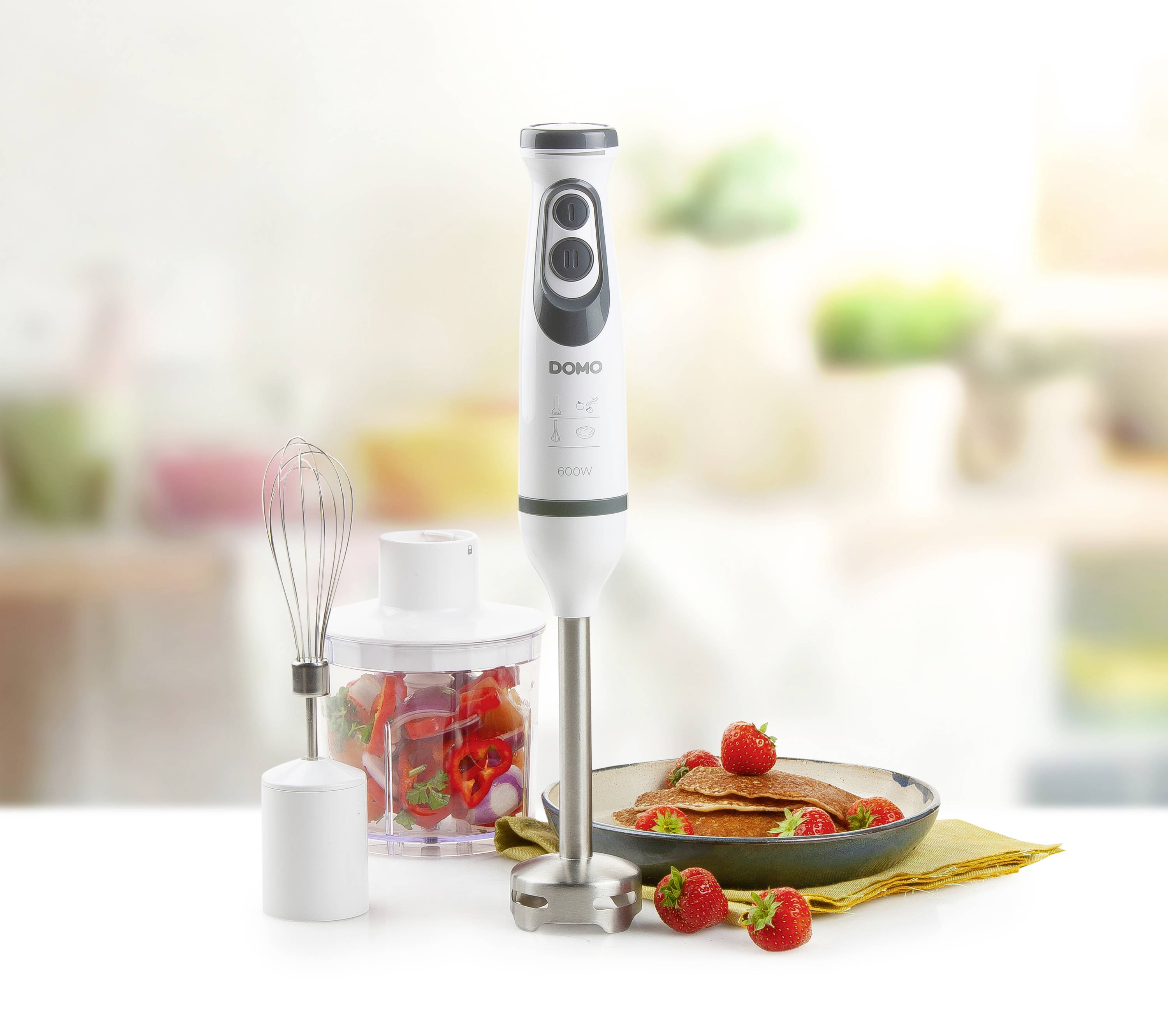 Ein Stabmixer mit Aufsätzen steht auf einem Tisch. Daneben sind frische Erdbeeren, ein Rührbesenaufsatz und ein gefülltes Gefäß zu sehen.