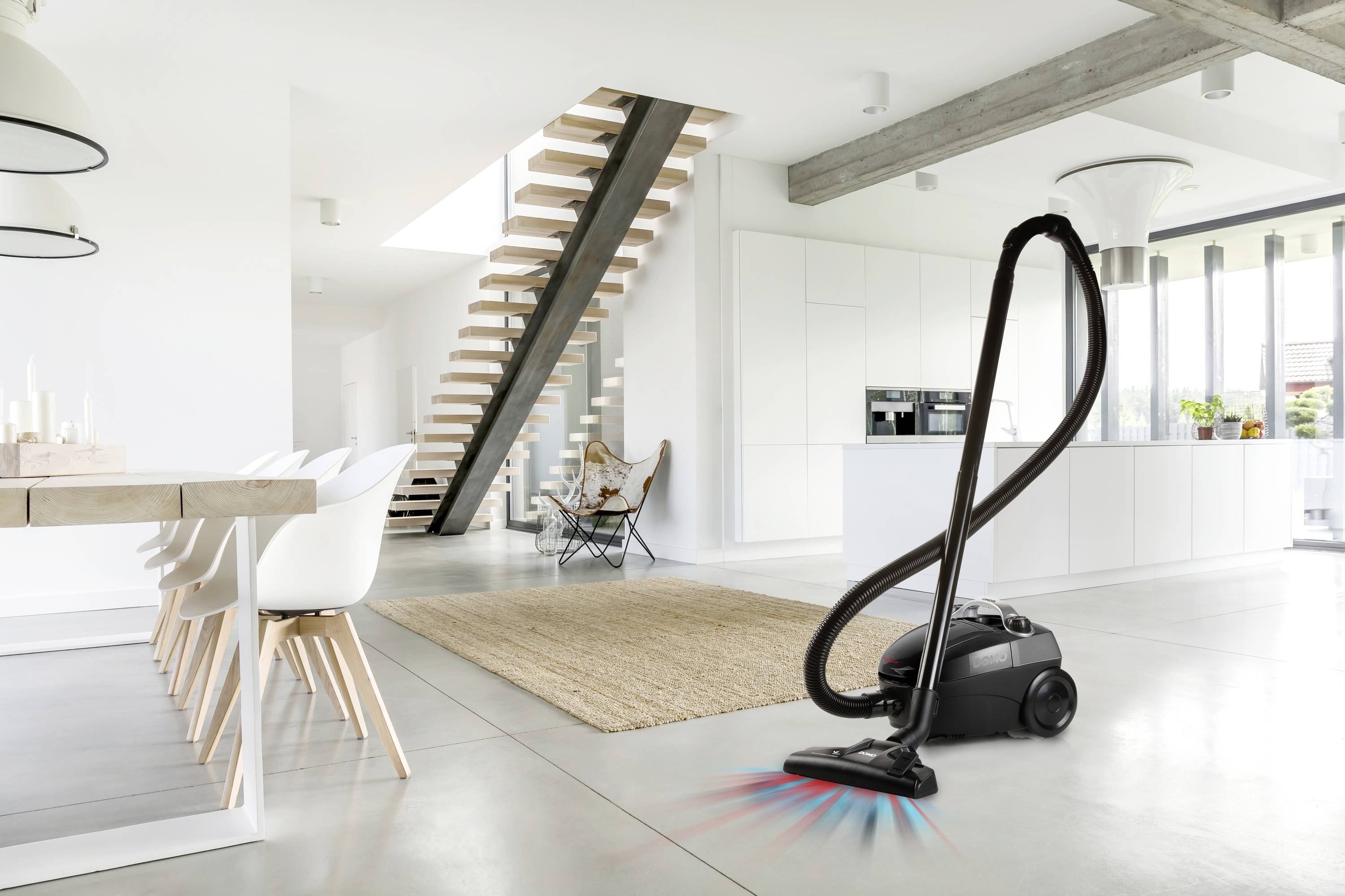 Ein Staubsauger steht in einem modernen Wohnzimmer mit weißen Möbeln und großer Treppe, bereit zum Reinigen des Bodens.