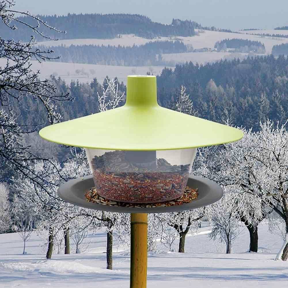 Ein grünes Vogelhäuschen mit Futter, im verschneiten Waldhintergrund. Keine sichtbaren Vögel. Winterschneelandschaft.