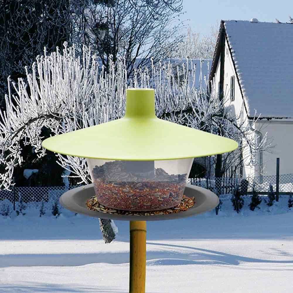 Ein Vogelhäuschen im verschneiten Garten, gefüllt mit Vogelfutter, vor einem Haus und frostbedeckten Bäumen.