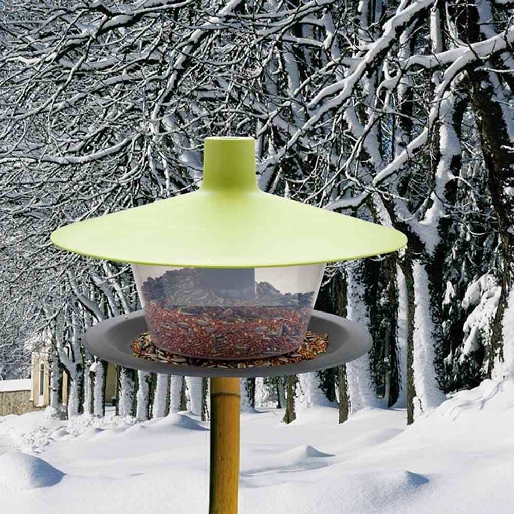 Ein Vogelhäuschen mit grüner Abdeckung und Samen gefüllt, steht in einem verschneiten Winterwald mit schneebedeckten Bäumen im Hintergrund.