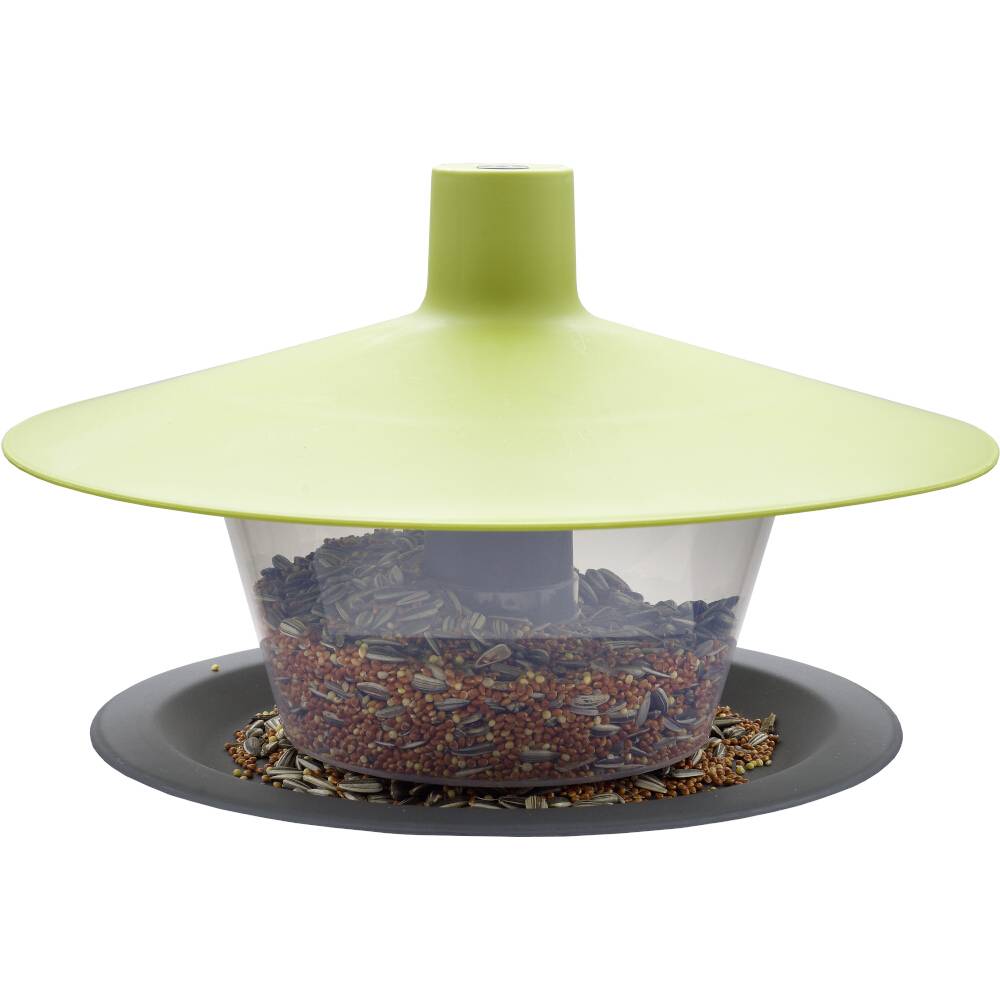 Ein grünes Vogelfutterhaus mit deckelartigen Dach, gefüllt mit verschiedenen Samen, auf einem kreisförmigen grauen Teller.