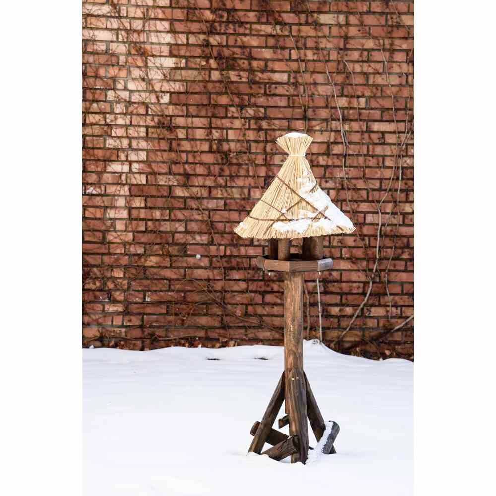 Vogelhaus mit Strohdach auf Holzbein in schneebedecktem Hof vor ziegelroter Mauer.