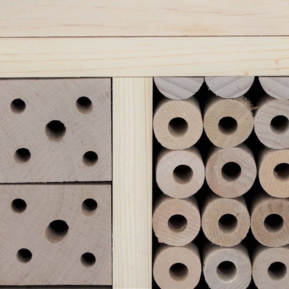 Holzbienenhaus aus hellem Holz mit zwei Bautypen: Links quadratische Blöcke mit Bohrlöchern, rechts runde Röhrenbündel.