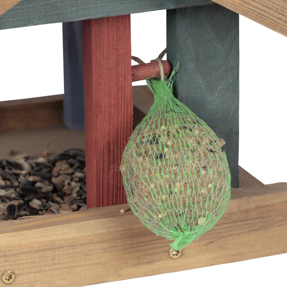 Ein Futterhäuschen mit einem grünen Netz voller Vogelfutterkugeln hängt daneben. Darunter liegen verschiedene Samen und Körner.