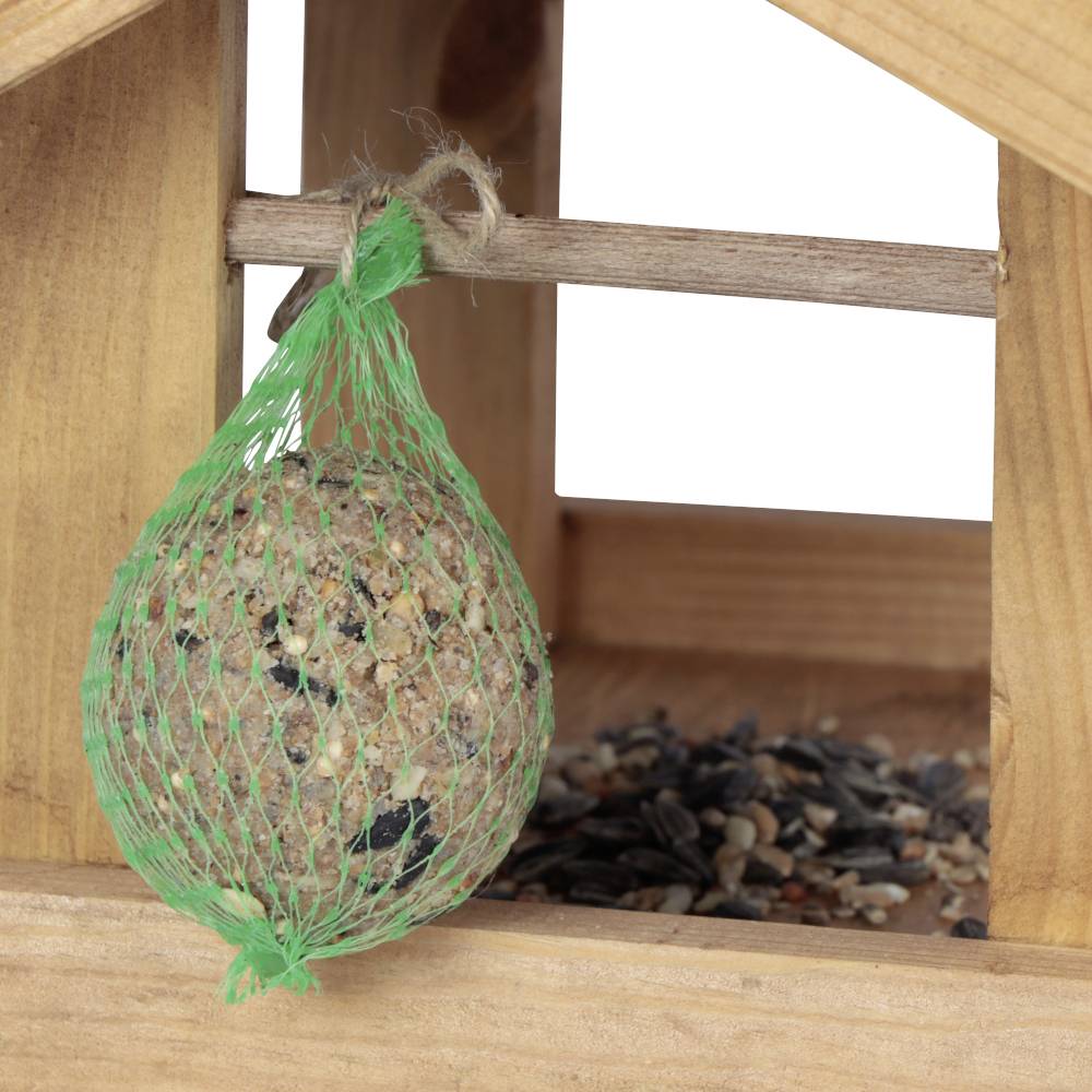 Ein Vogelhäuschen aus Holz mit einem Meisenknödel in einem grünen Netz. Im Hintergrund sind lose körner- und Samenmischungen zu sehen.