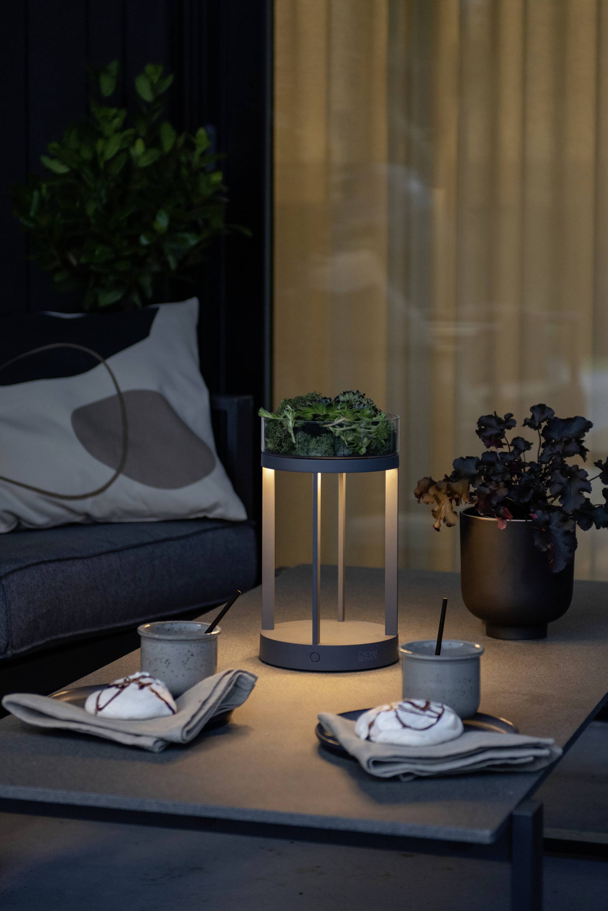 Gedeckter Tisch auf Terrasse mit Tischlampe, zwei Dessertteller mit Sahnehaube und Schokoladensauce, Pflanze im Hintergrund.