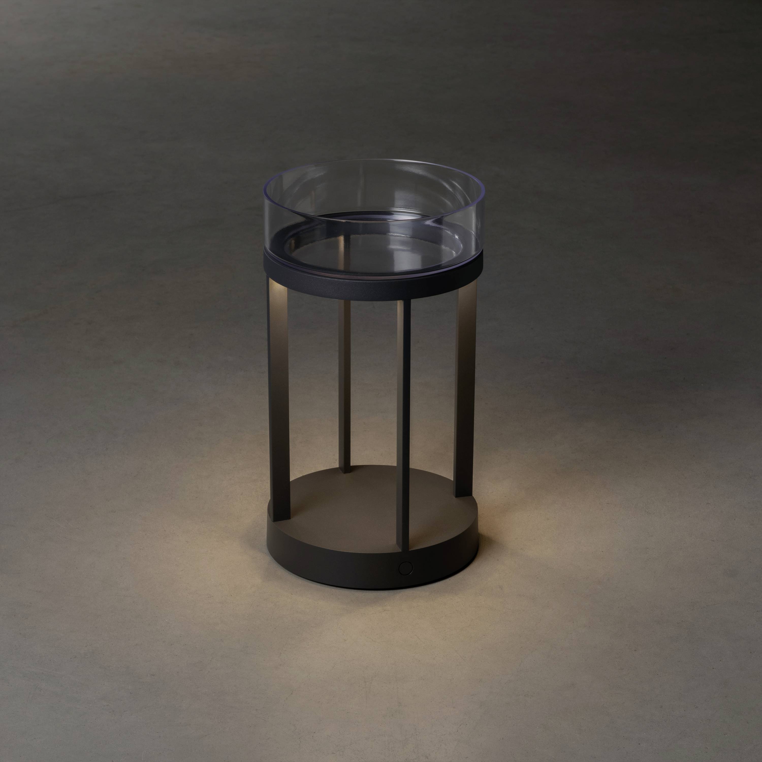 Ein kleiner, runder Nachttisch mit Glasplatte und dunklem Metallgestell steht auf einem neutralen Boden.