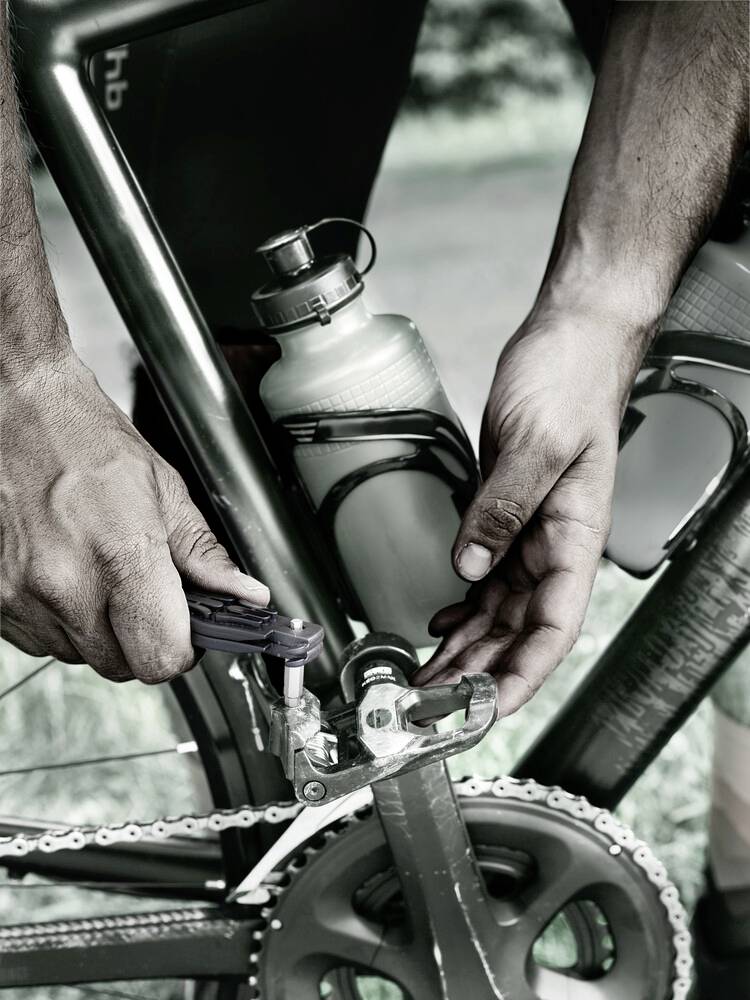 Person repariert Fahrradgetriebe mit Werkzeug; Wasserflasche am Rahmen befestigt; Fokus auf Hände und Pedal.