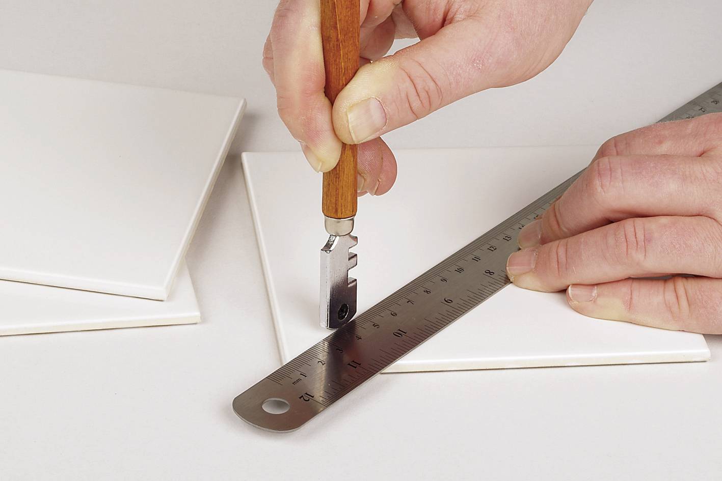 Eine Person schneidet mit einem Fliesenschneider und einem Metalllineal eine weiße Fliese.