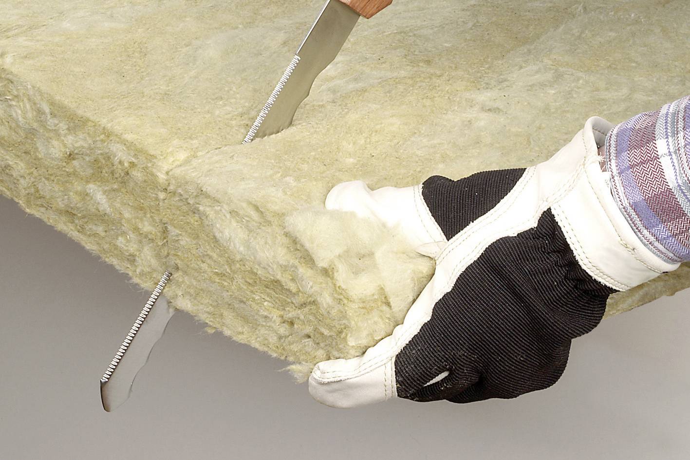 Eine Person schneidet mit einem Messer eine Dämmplatte aus Mineralwolle. Hände sind mit Arbeitshandschuhen geschützt.