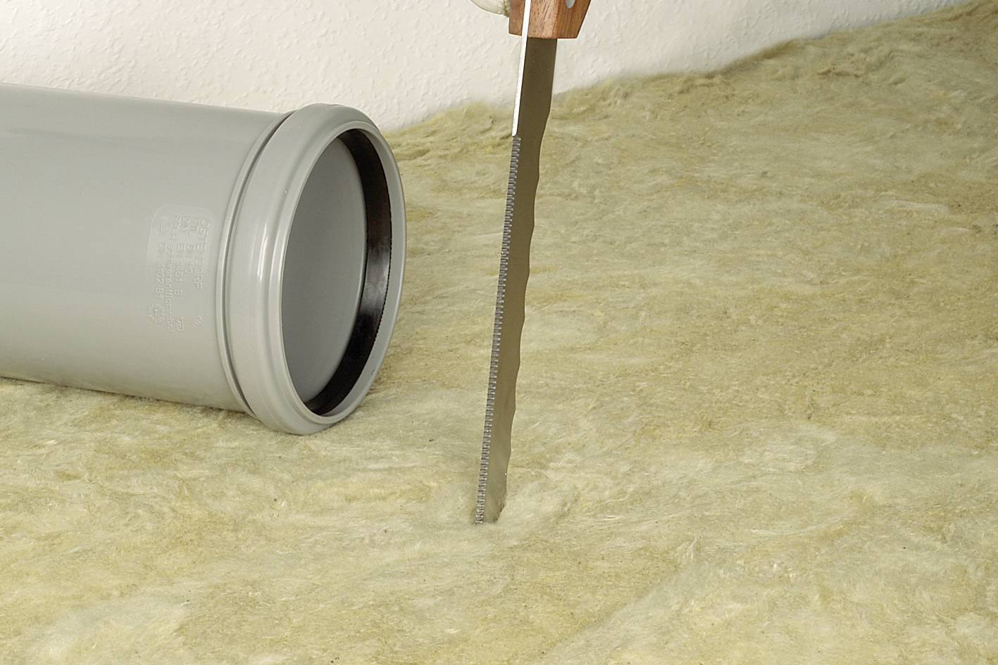 Eine Säge schneidet durch eine Matte aus Isoliermaterial auf einem Boden, neben einem grauen Plastikrohr liegend.