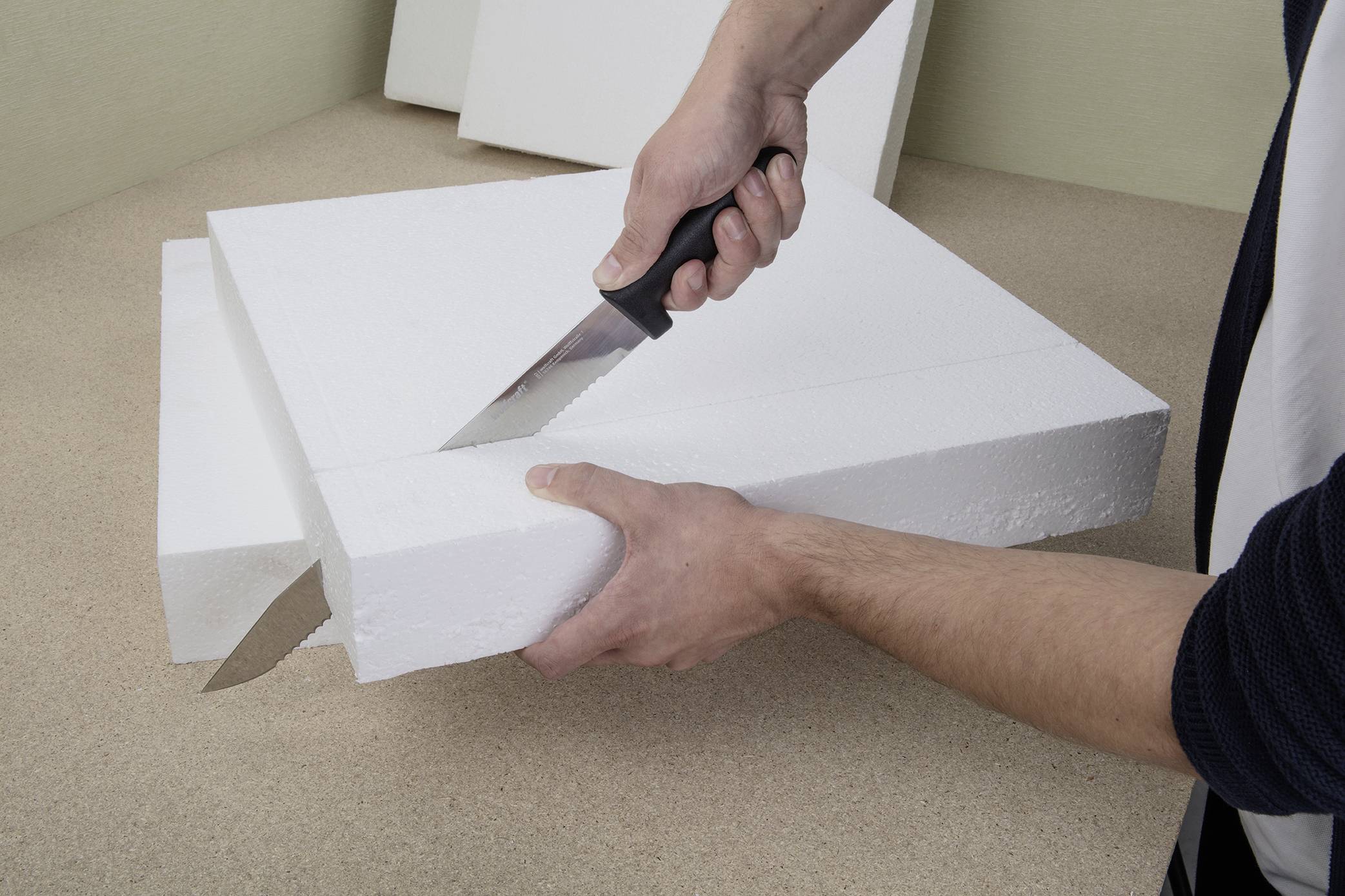 Eine Person schneidet einen großen Styroporblock mit einem Messer. Der Untergrund ist ein Tisch oder eine Arbeitsfläche.