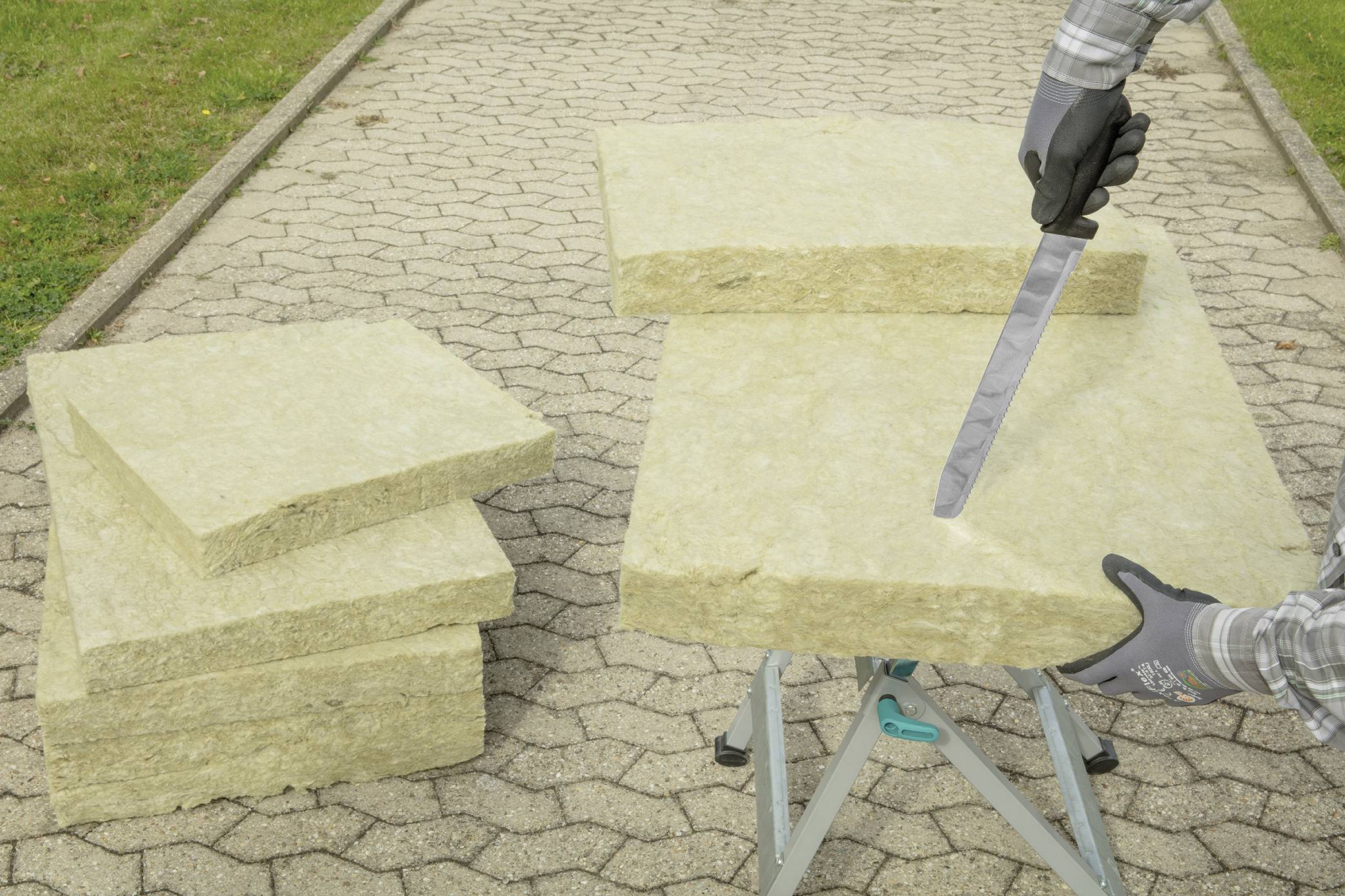 Eine Person schneidet Mineralwolle-Dämmplatten mit einem Messer auf einem Klappbock im Freien. Weitere Platten liegen gestapelt daneben.
