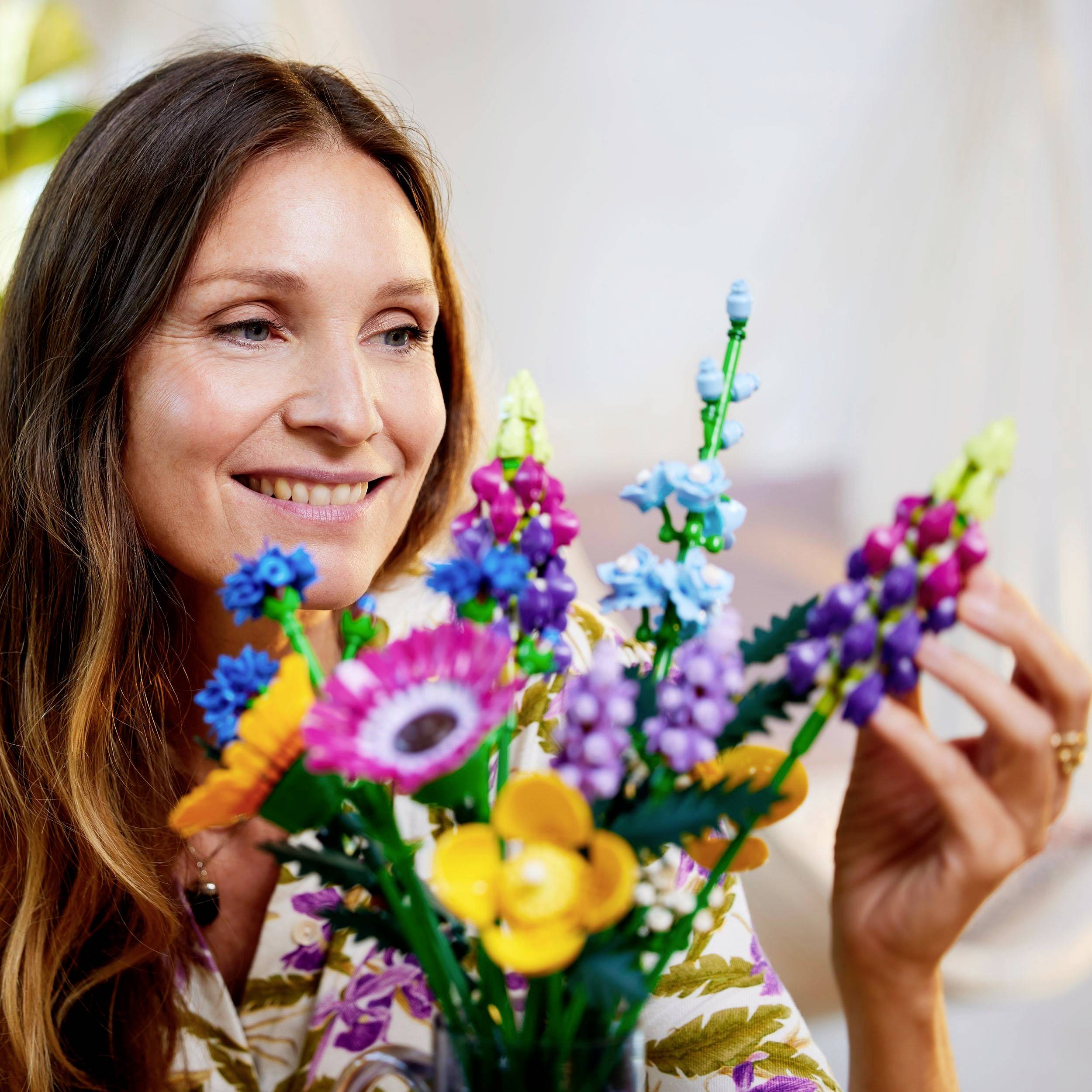 Eine Frau lächelt, während sie einen bunten Blumenstrauß aus LEGO-Steinen betrachtet. Im Hintergrund sind unscharfe Pflanzen zu sehen.