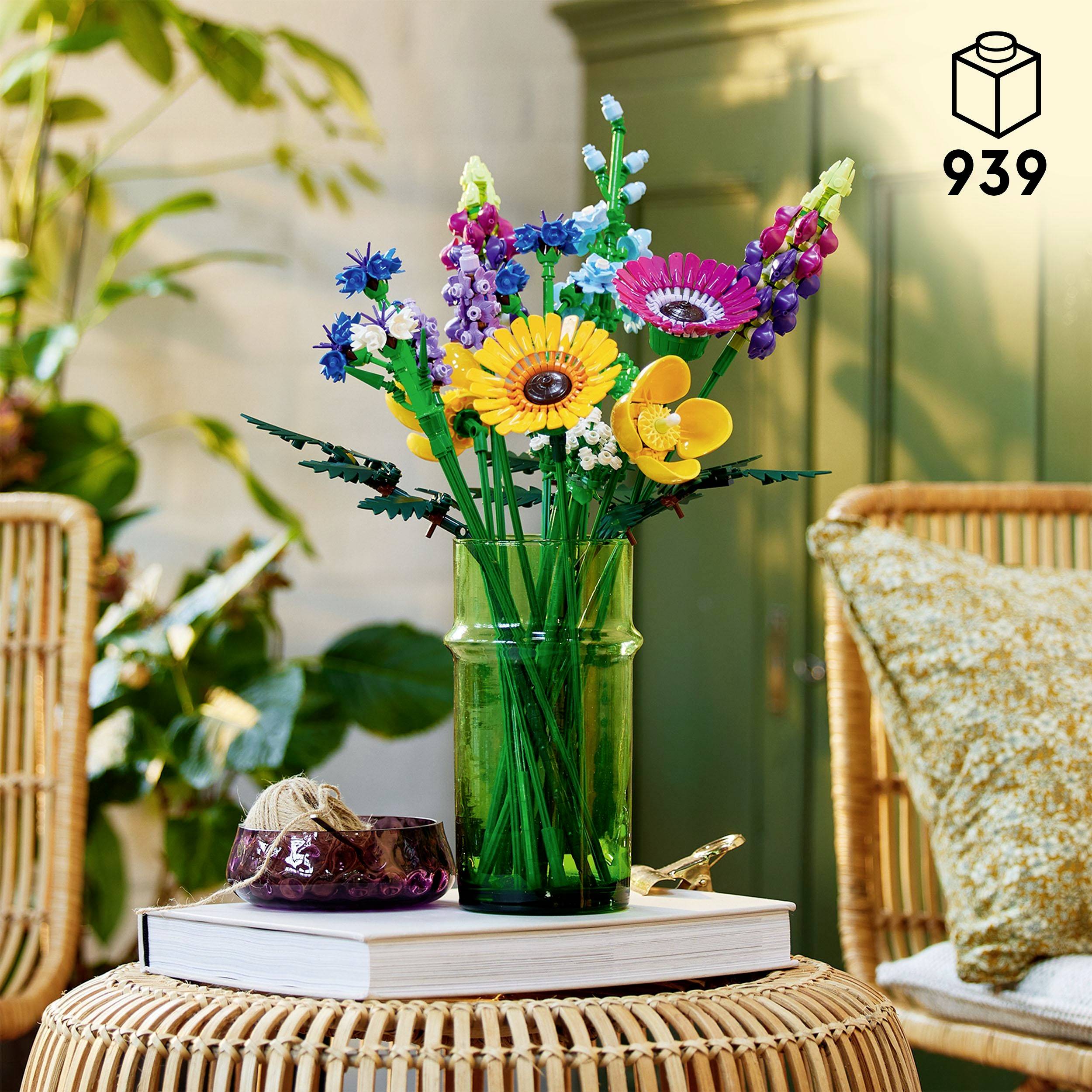Ein grüner Glasvase mit bunten LEGO Blumen steht auf einem weißen Tisch. Im Hintergrund sind Topfpflanzen und ein Möbelstück sichtbar.