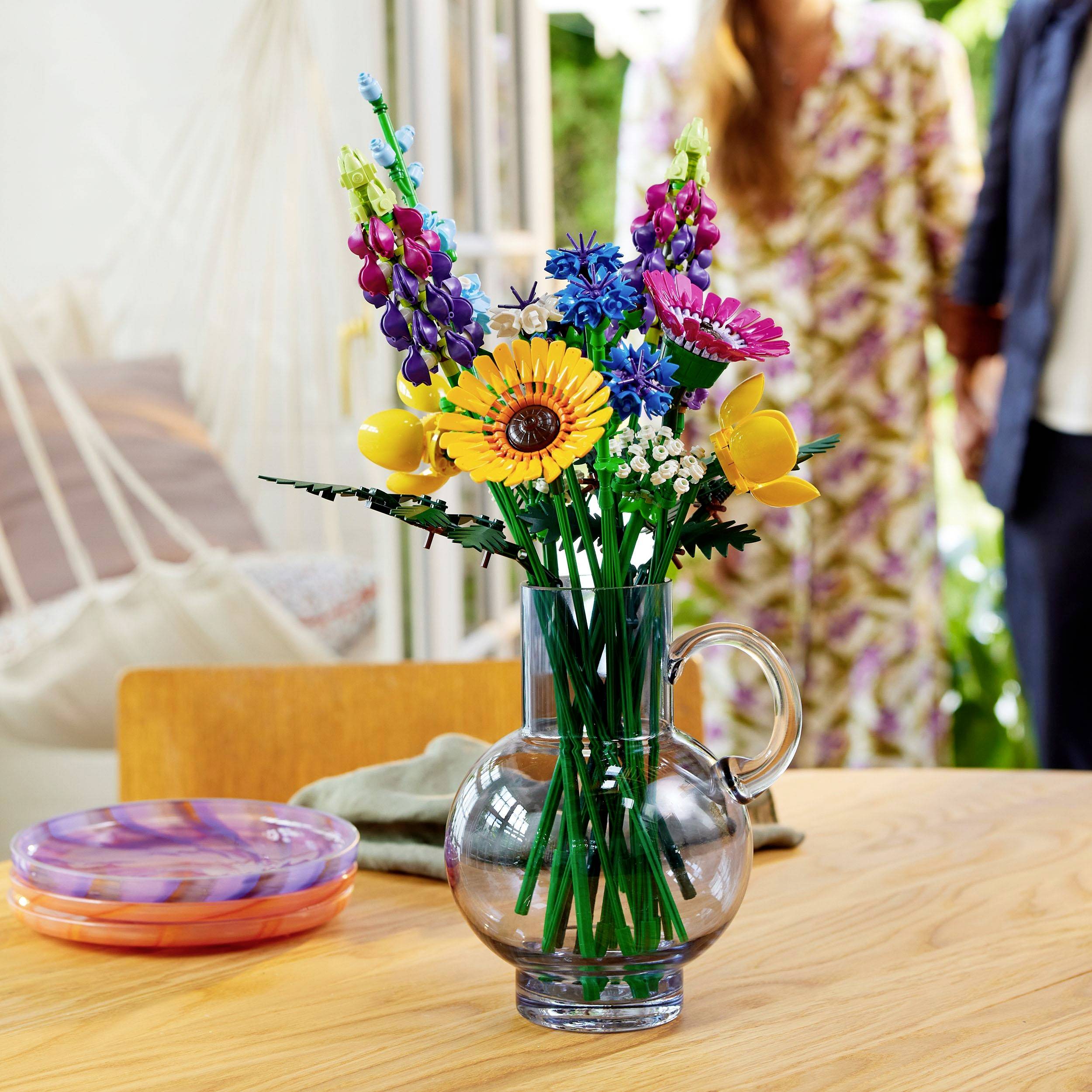 Ein bunter Strauß aus gemischten Blumen in einer klaren Vase steht auf einem Holztisch, im Hintergrund sind zwei Personen und ein offenes Fenster sichtbar.