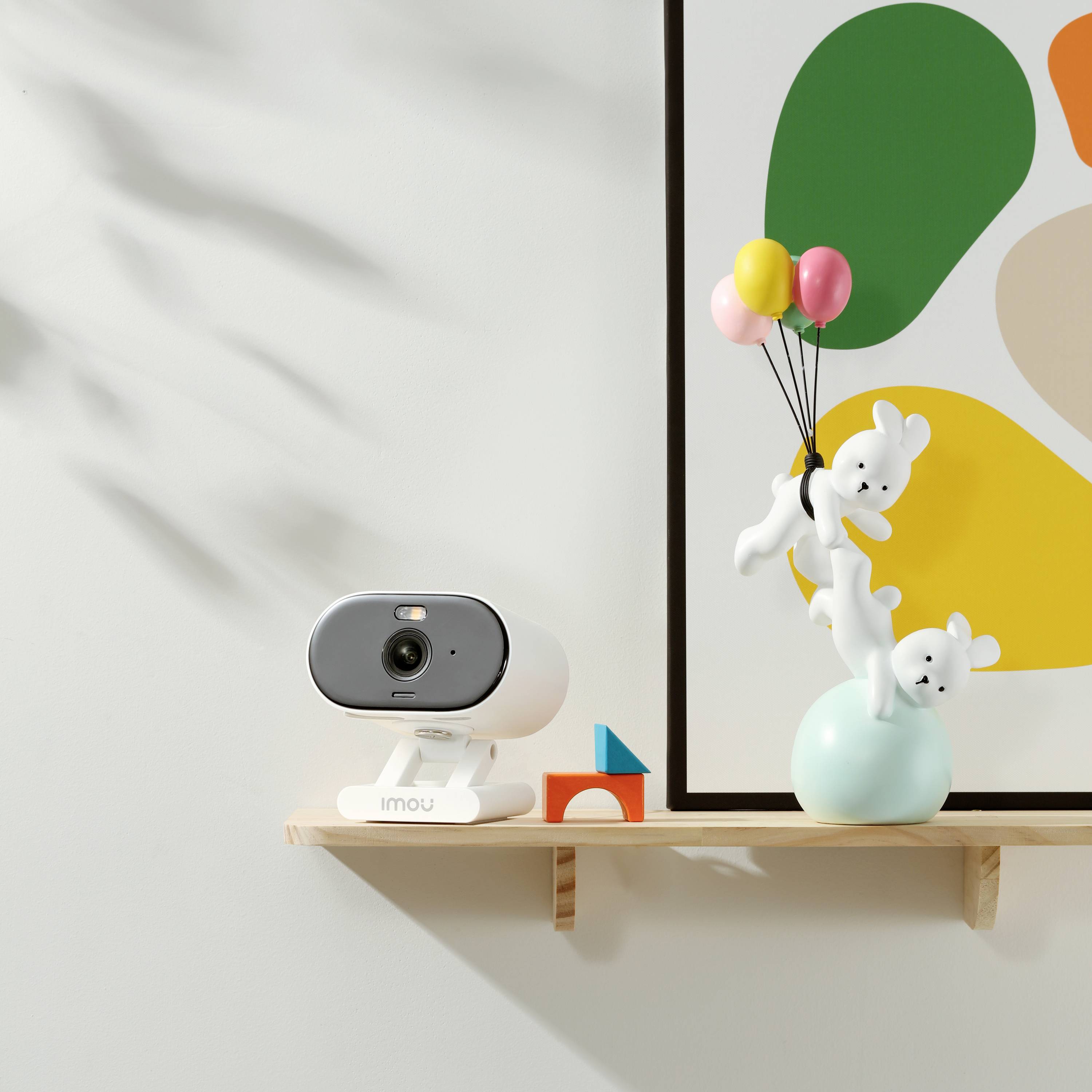 Weiße Überwachungskamera auf Holzregal neben bunter Grafik. Vase mit Luftballons und Stofftieren, bunte Bauklötze daneben.