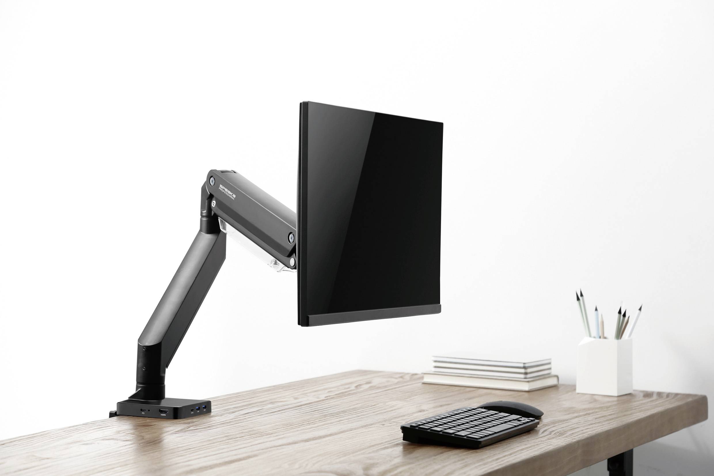 Ein Computermonitor ist an einem flexiblen Halterarm über einem Schreibtisch montiert. Auf dem Tisch liegen Notizbücher und Stifte.