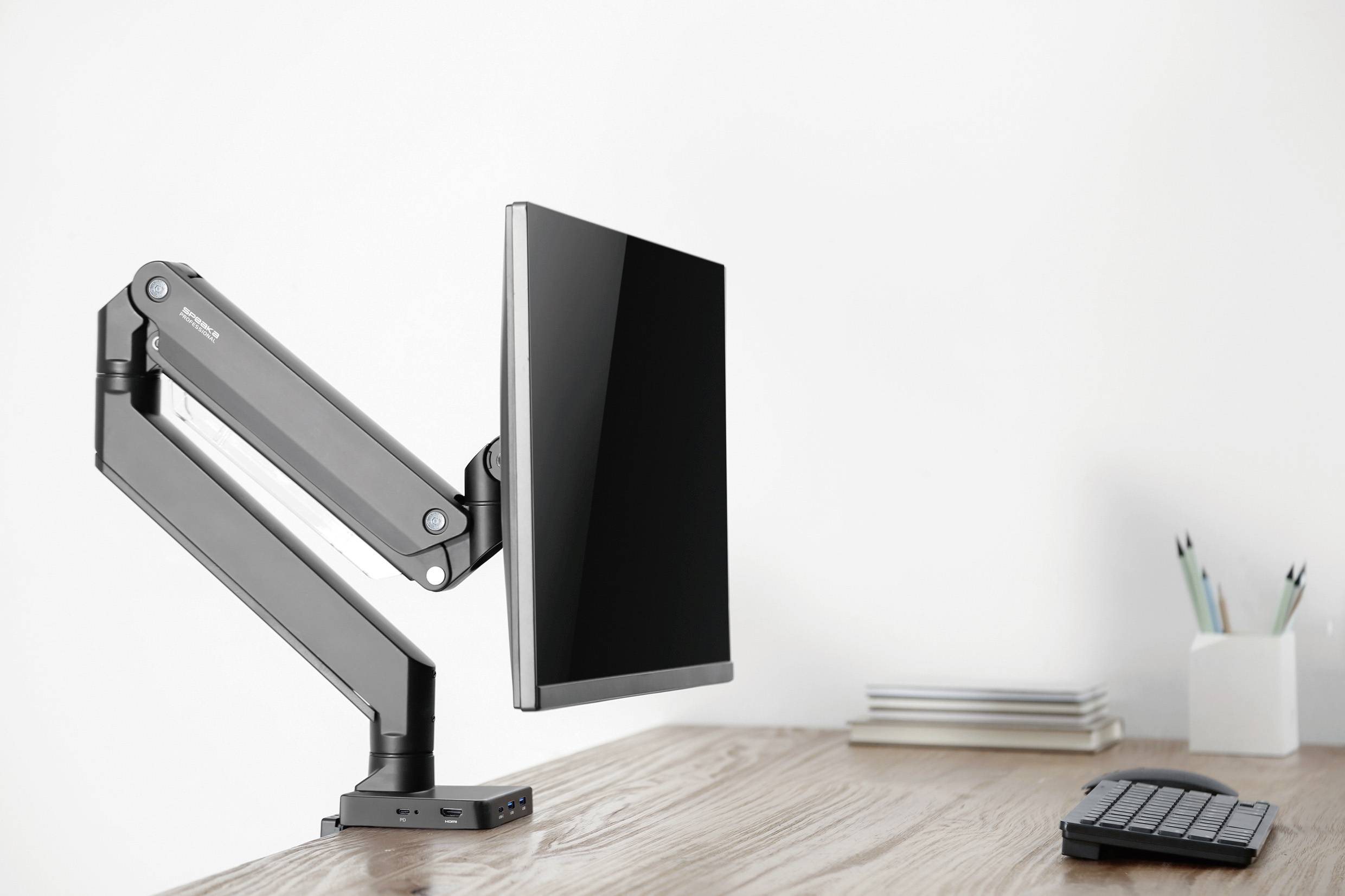 Monitorarm mit montiertem Bildschirm auf einem Holztisch, neben einer Tastatur und Schreibwerkzeugen; minimalistische Büroeinrichtung.