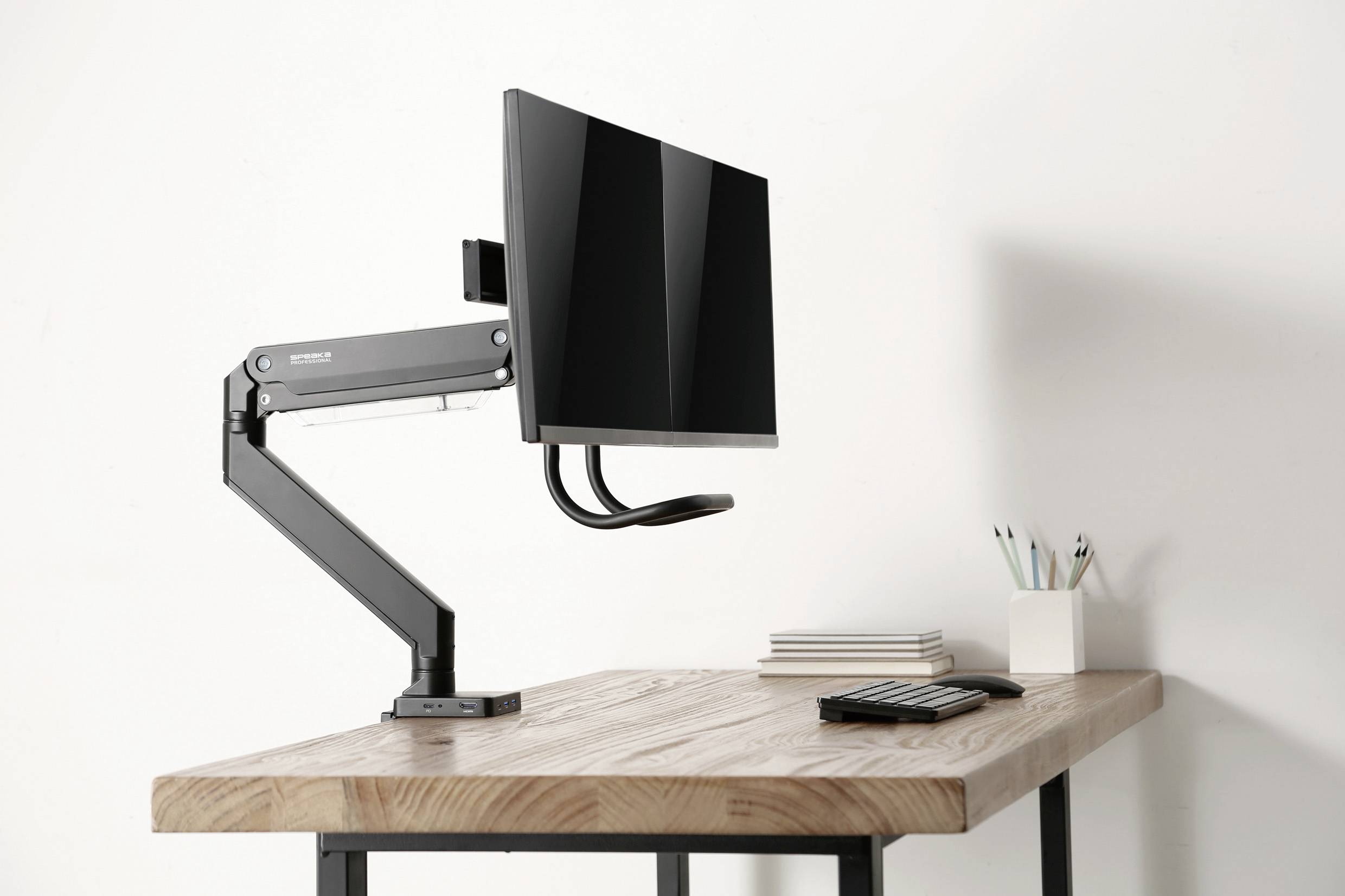 Der Schreibtisch zeigt einen modernen, einstellbaren Monitorarm mit einem montierten Bildschirm. Daneben liegen Stifte, Notizbücher und ein Taschenrechner.
